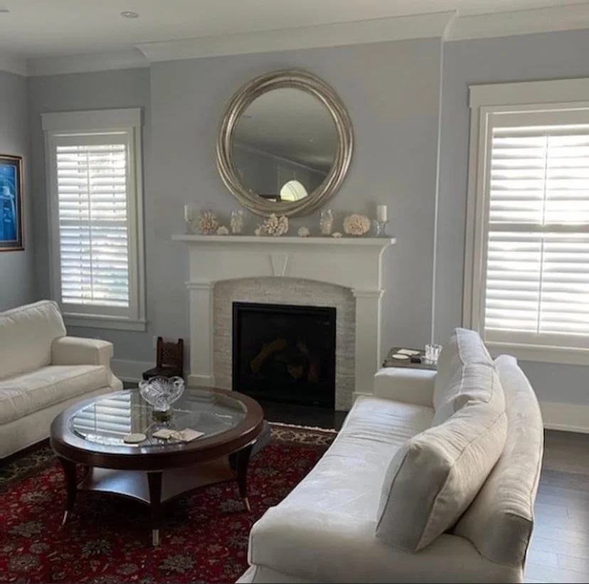 A living room with two cream-colored sofas, a wooden coffee table, a fireplace with a round mirror above it, and two windows with white plantation shutters.