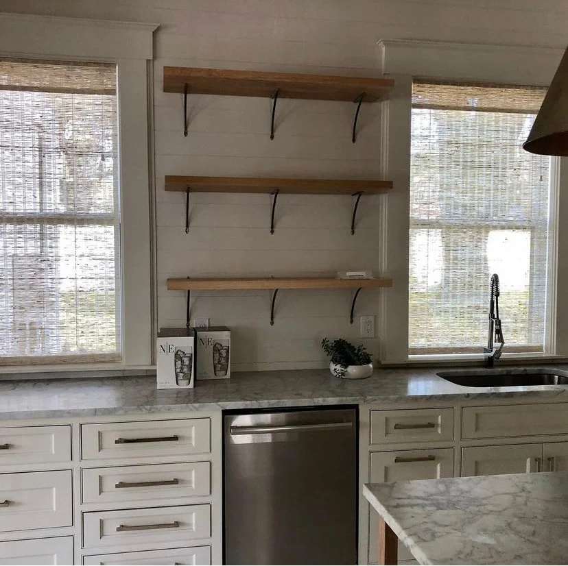 Kitchen with white cabinets, marble countertops, two large windows with woven shades, open shelves on the wall, a sink with a black faucet, a small potted plant, and two magazines on the counter.
