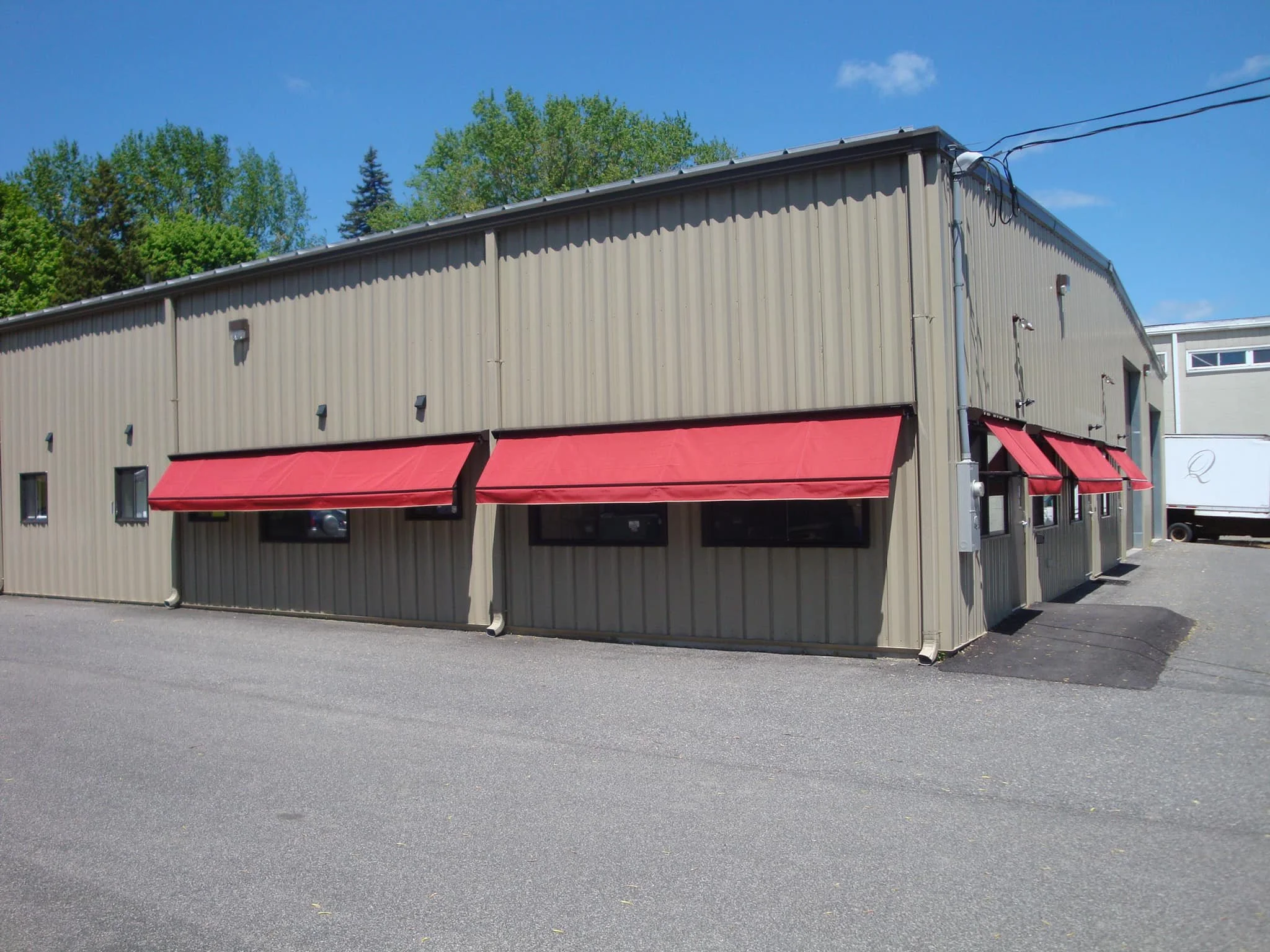 A beige commercial building with three red awnings and four small windows, located on a paved lot under a partly cloudy sky.