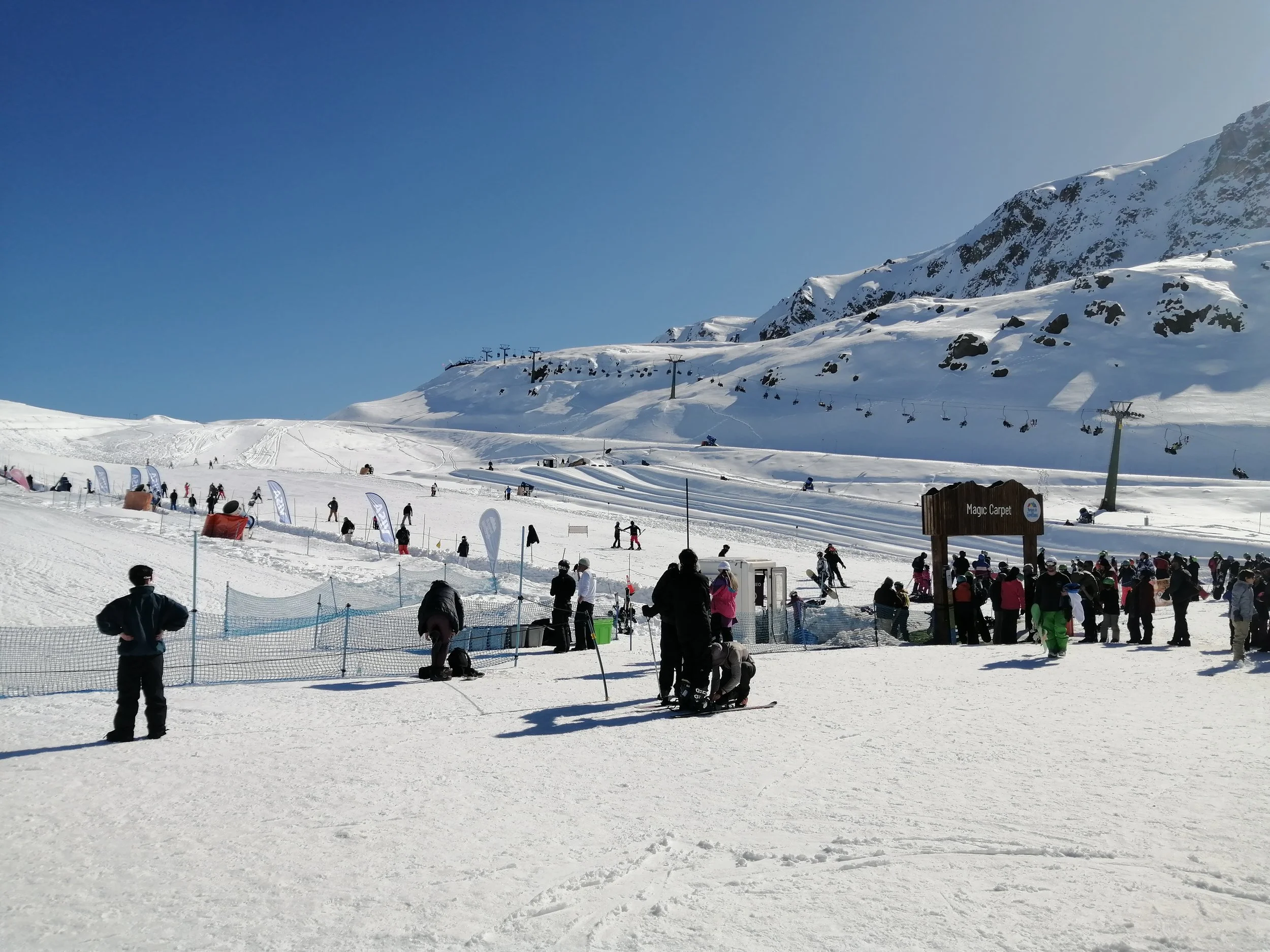 A busy snow-covered ski resort with skiers and snowboarders enjoying the slopes, a ski lift in operation, and a mountain in the background.