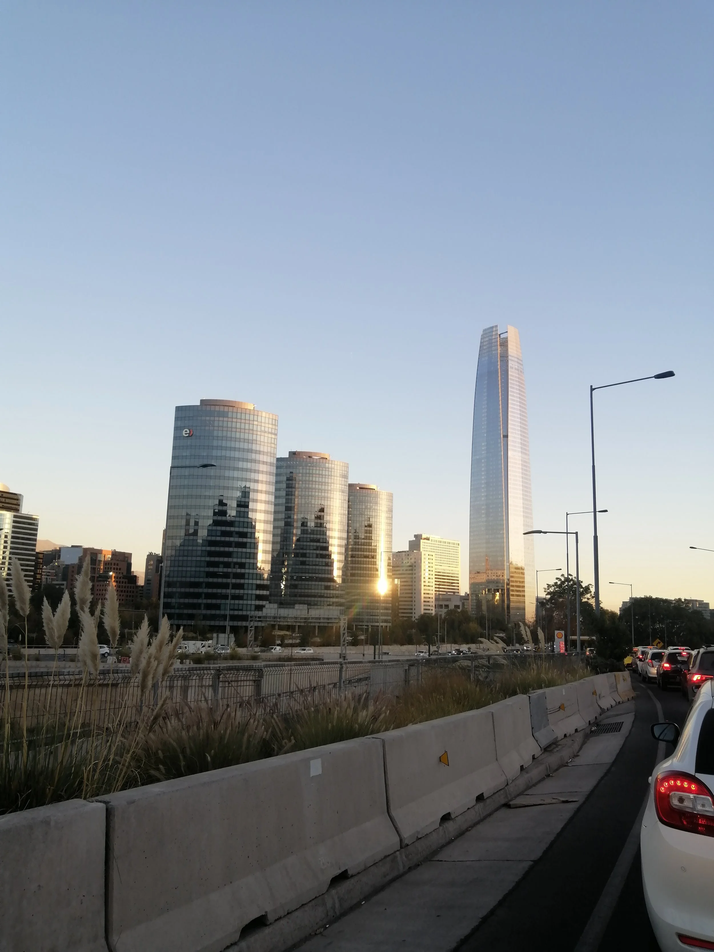 City skyline with tall glass skyscraper reflecting sunlight, several shorter buildings, streetlights, and cars on a busy road at sunset.