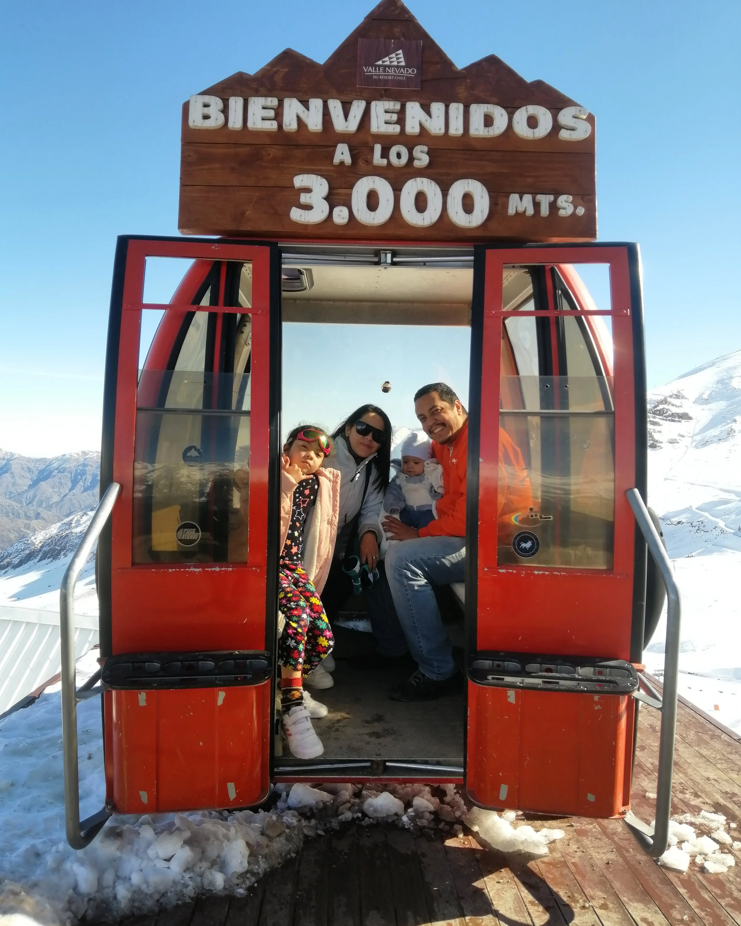 A group of people standing inside a red gondola with a wooden sign above it, welcoming visitors at 3,000 meters elevation. The sign reads 'Bienvenidos a los 3,000 mts.' and is set against a backdrop of snowy mountains and a clear blue sky.