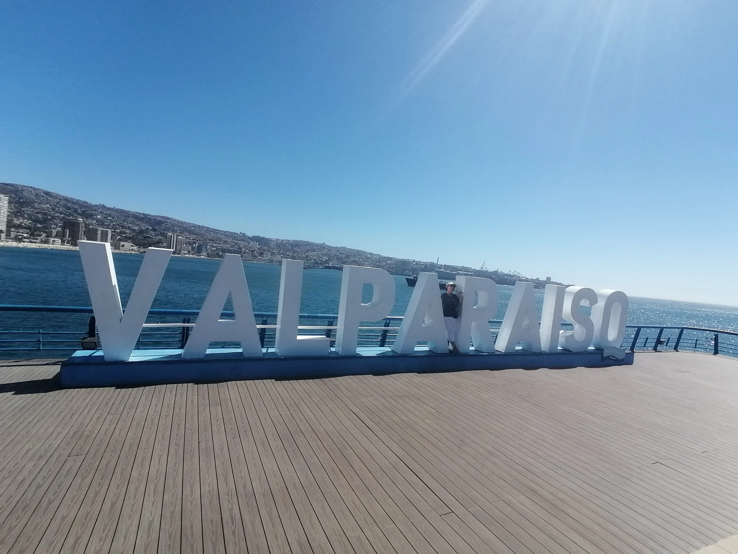 Clear blue sky over San Francisco Bay with a city skyline on the hillside and large white letters spelling 'WASHINGTON' on a wooden deck, with a person standing near the letters.