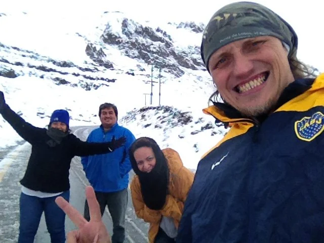 Four people smiling and posing outdoors in a snowy mountainous area, one making a peace sign with their hand.