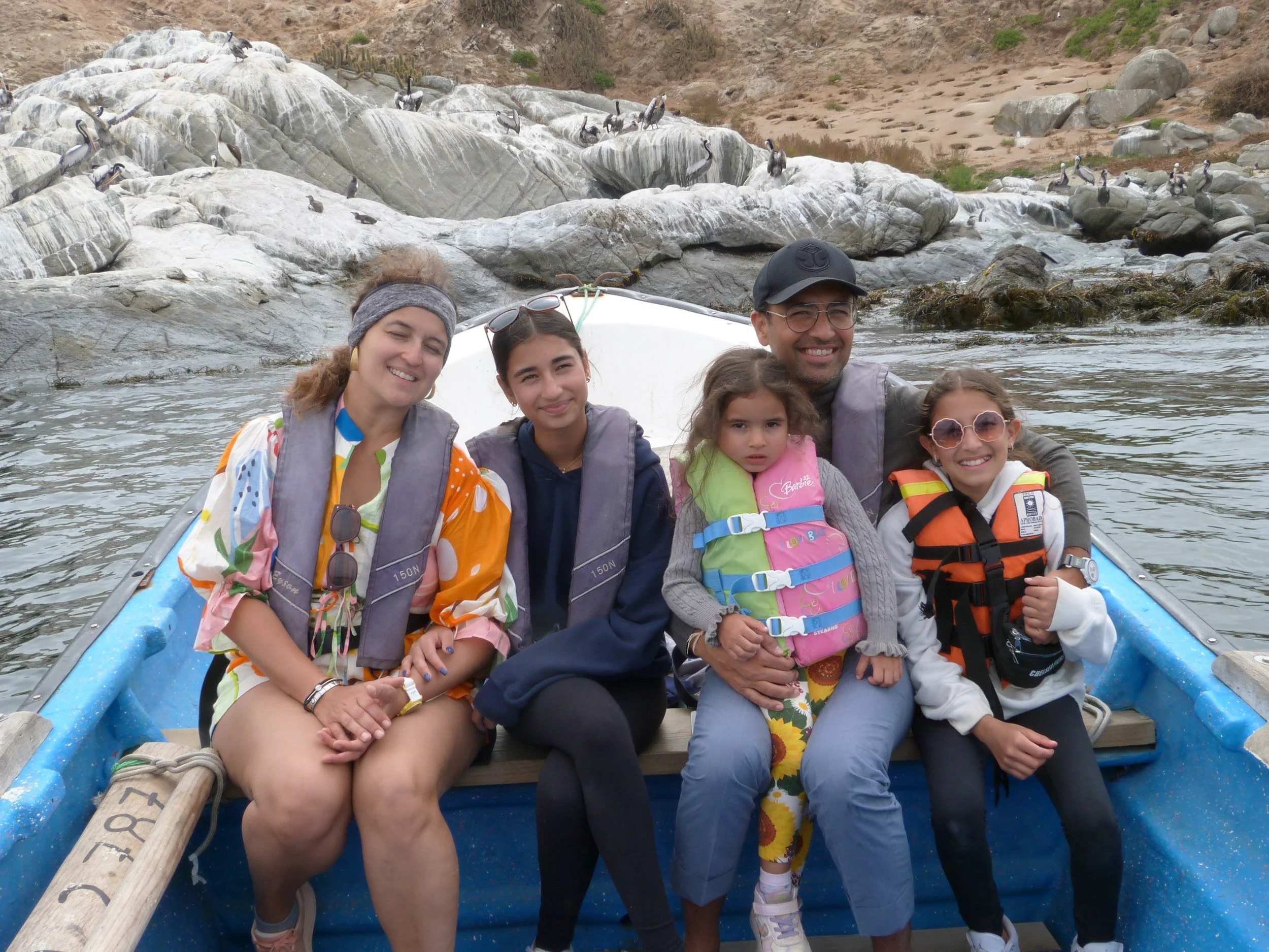 Group of five children and one adult smiling and sitting in a blue boat on a river, with rocky terrain and pelicans perched on rocks in the background.