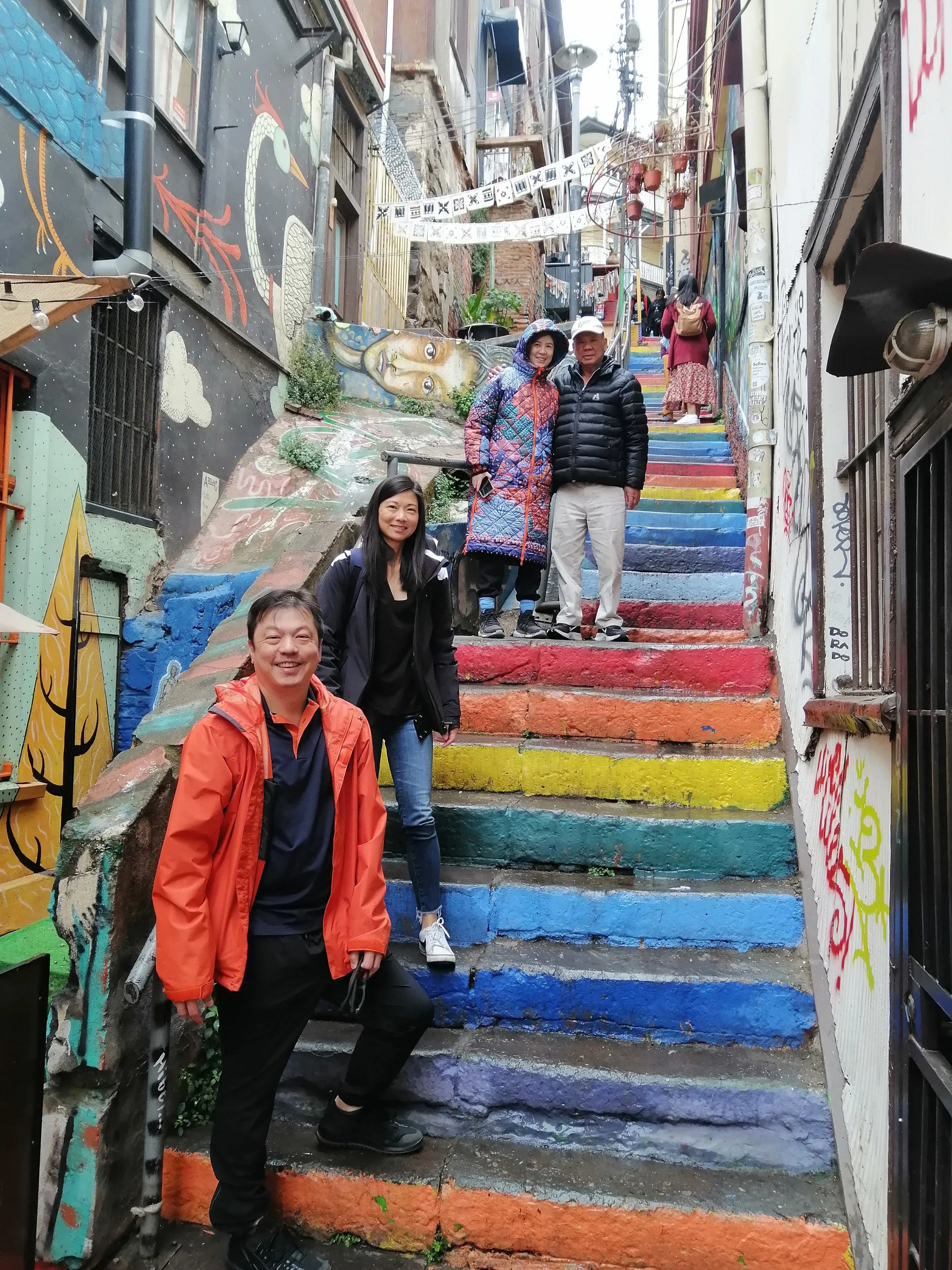 A group of four people, three women and one man, are standing on colorful painted steps in an alleyway. The steps are painted in bright rainbow colors. The alley features murals and graffiti on the walls, with one wall showing a dragon and another a portrait of a woman. Some people are walking further up the stairs, and the scene has a lively, artistic atmosphere.