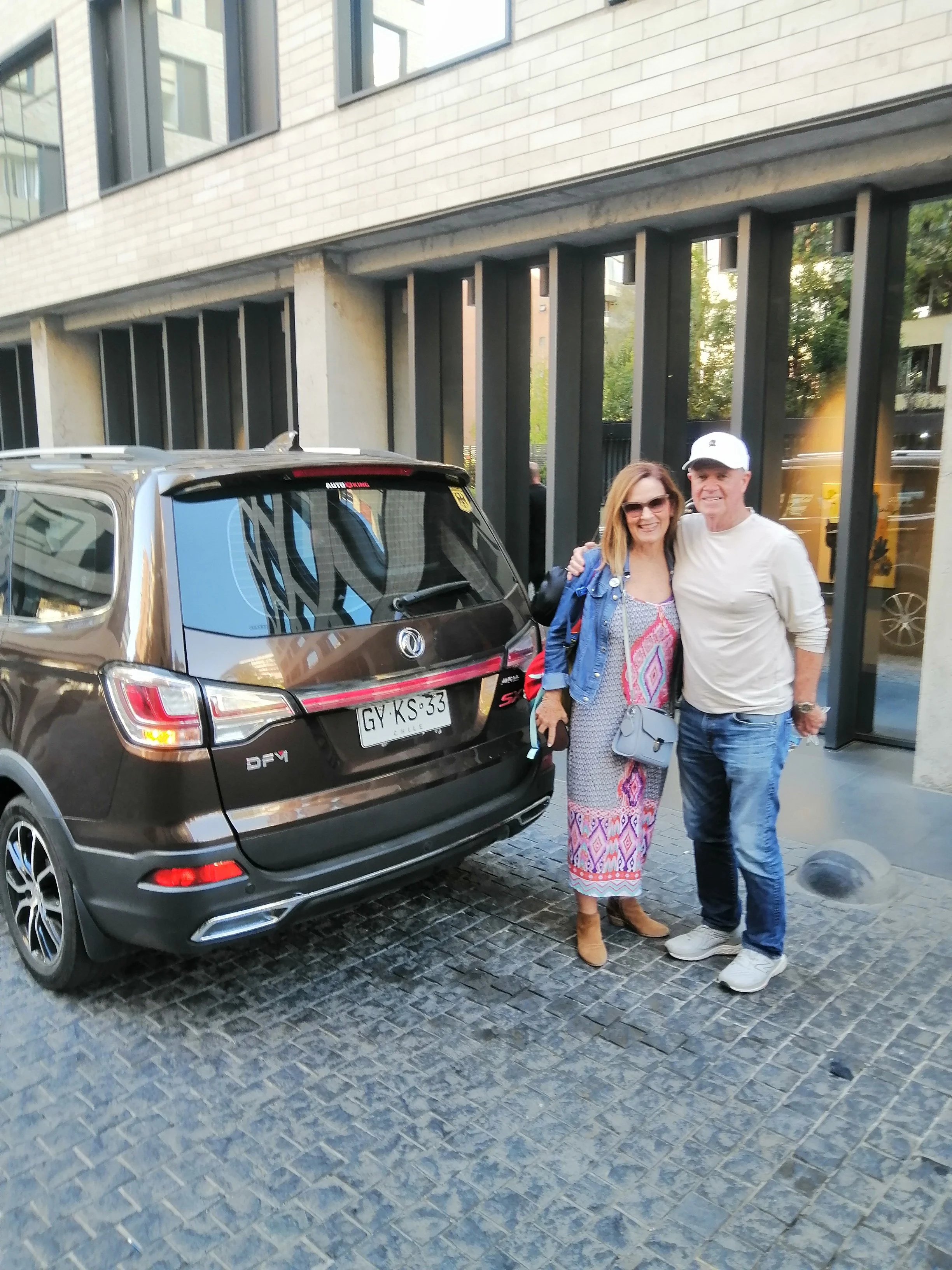 A woman and a man smiling and posing together on a cobblestone street next to a black SUV, with a building with large windows behind them.