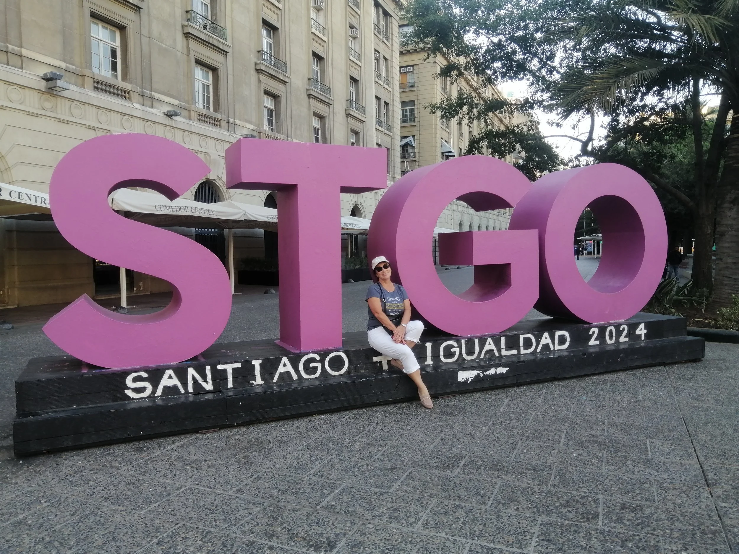A woman wearing sunglasses, a hat, a blue shirt, and white pants is sitting on a black platform in front of large pink letters spelling 'Santiago', with a building and trees in the background.