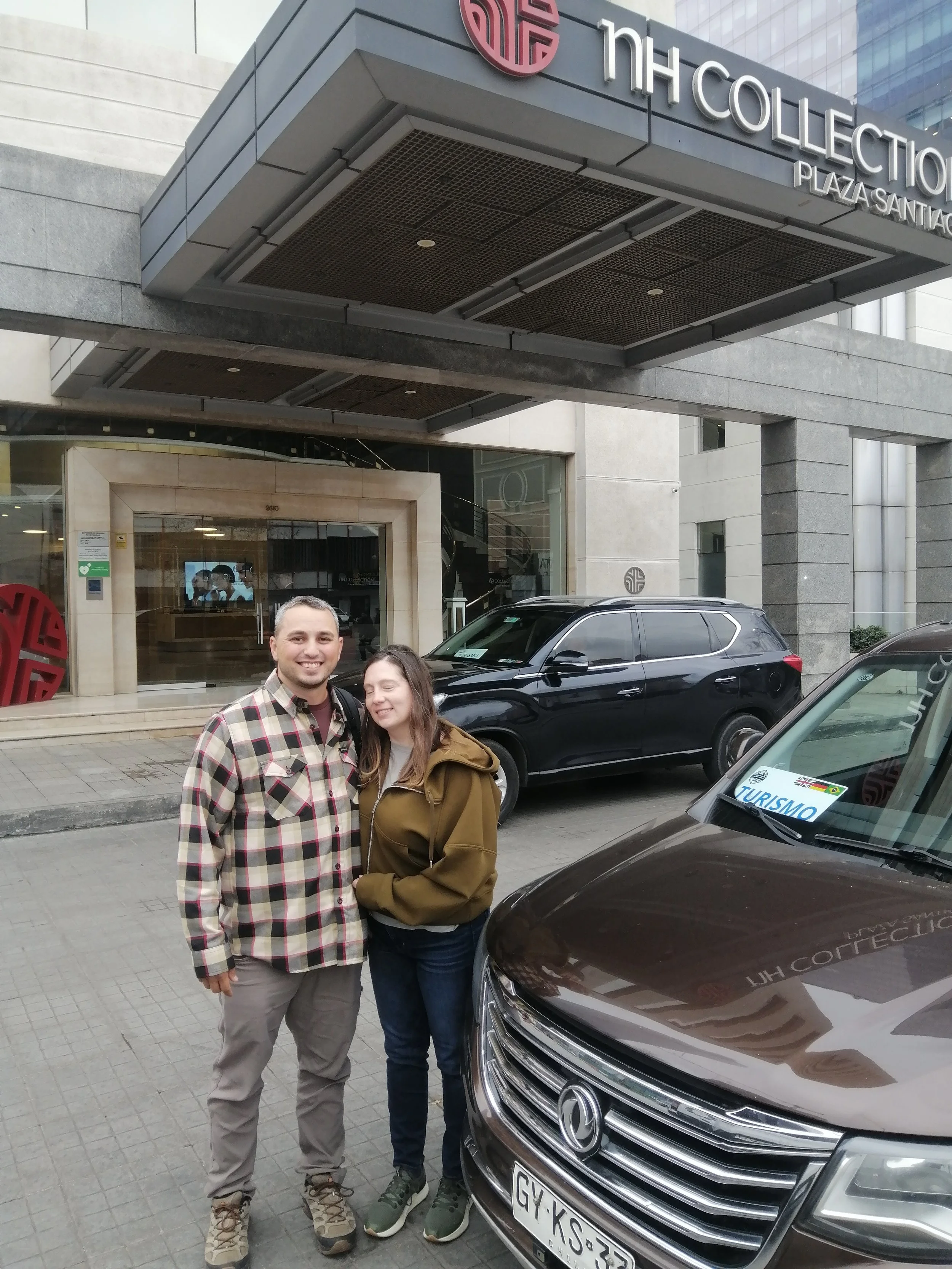 A smiling man and woman standing in front of a large gray car parked on the street, in front of a modern building with a sign that reads 'NH Collection Plaza Santiago.' The man is wearing a plaid shirt and khaki pants, the woman is wearing a brown jacket and dark pants. There are other cars and a building with glass windows in the background.