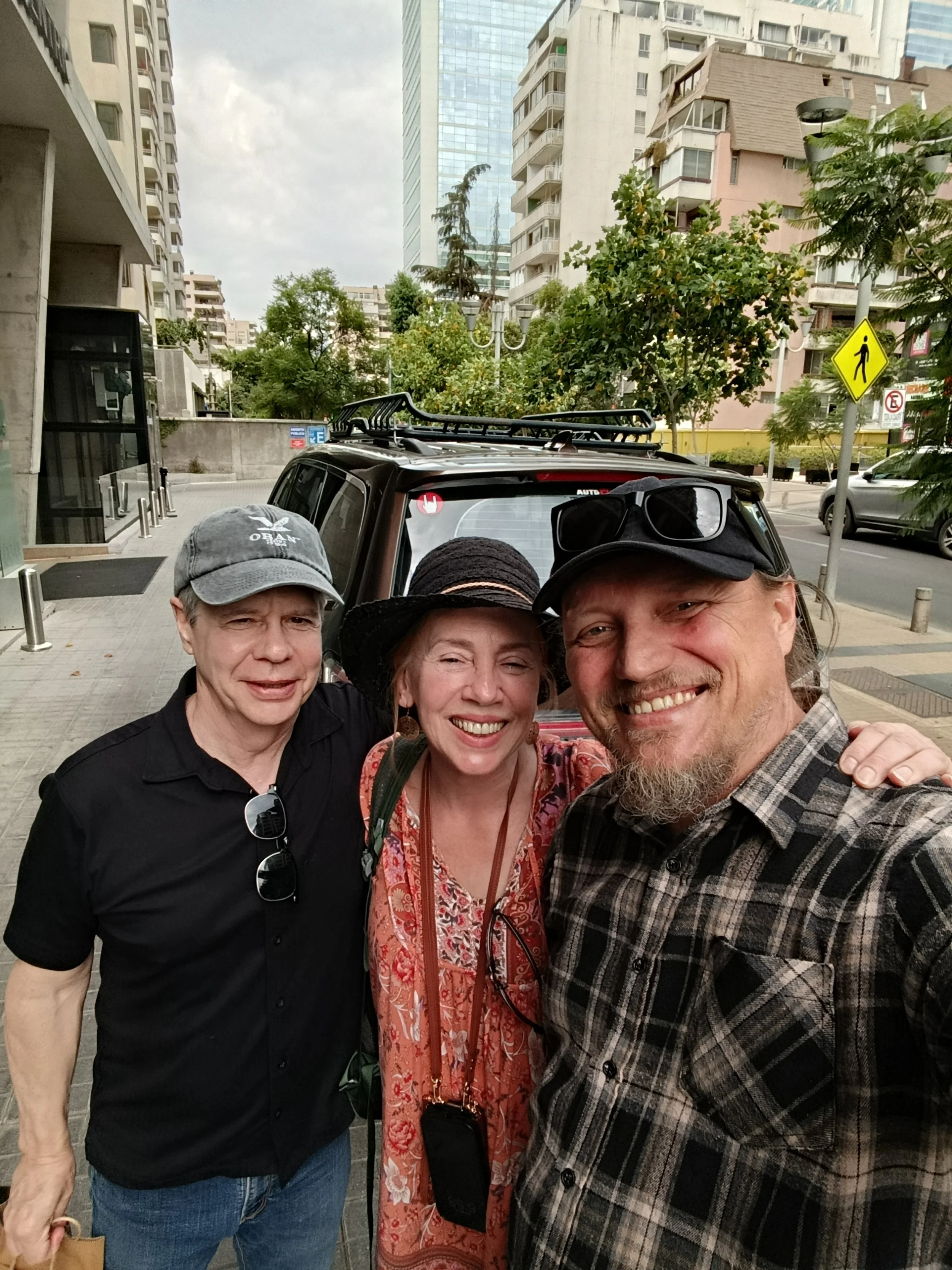 Group of three people taking a selfie on a city street, with tall buildings and trees in the background.