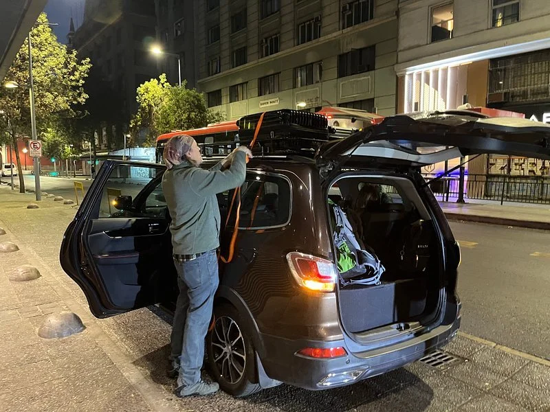 A man in a gray hoodie and jeans is loading equipment into a black SUV at night on a city street. The SUV's rear hatch is open, and the street is illuminated by streetlights, with buildings and trees visible in the background.