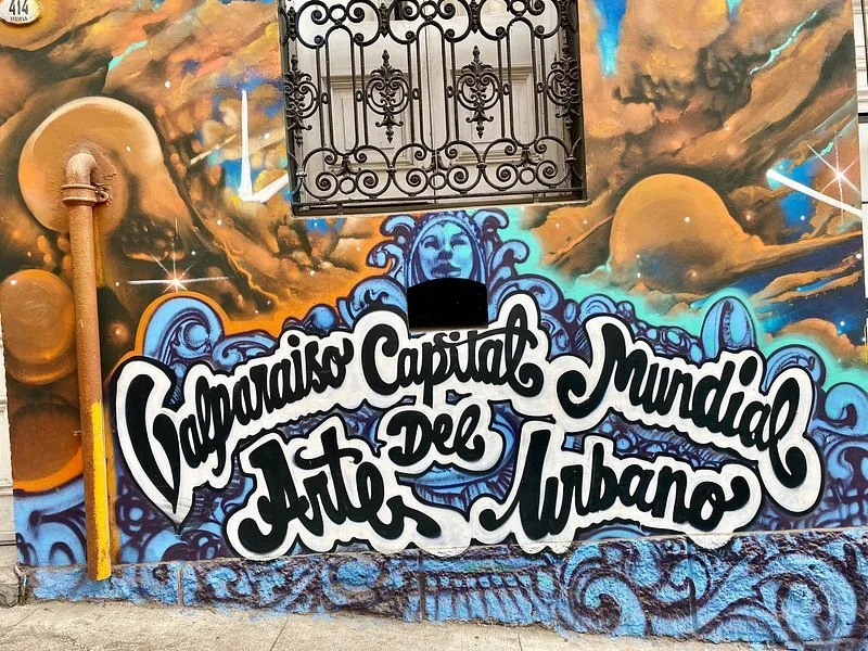 Colorful mural painting with clouds, planets, and artistic elements, featuring the text 'Galerías Capital del Arte Urbano' in bold black and white letters.