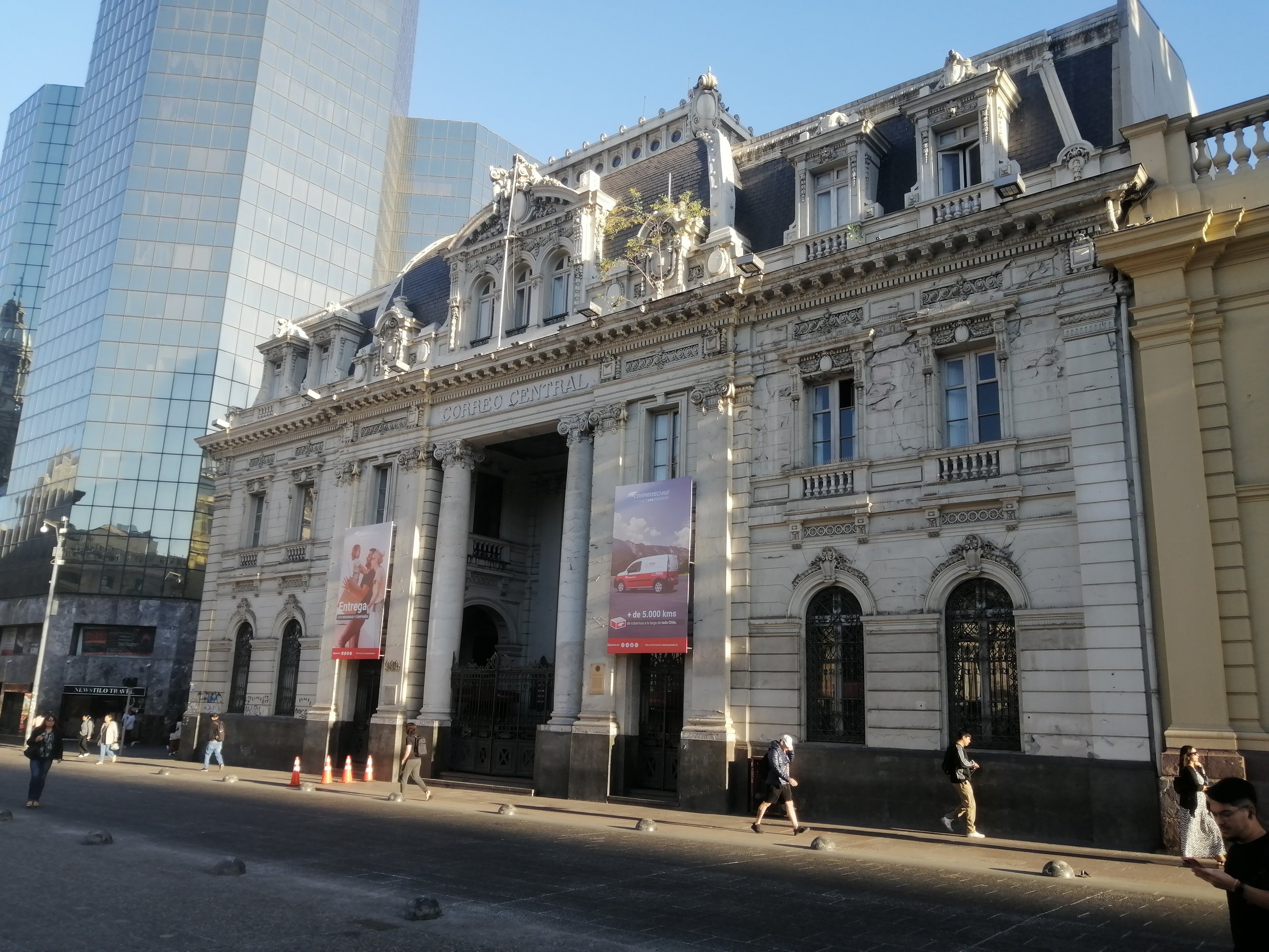 Historic building with ornate architecture and signage stating 'Correo Central,' modern glass skyscrapers in the background, people walking on the sidewalk, and banners hanging in front of the building.