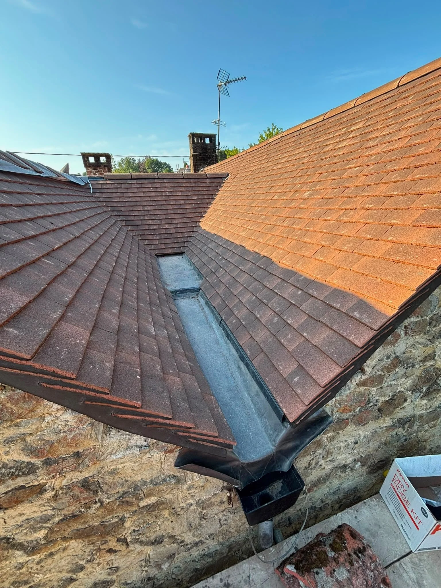 Close-up of a roof with reddish-brown tiles, a skylight window, a brick chimney, a TV antenna, and a black downpipe, with a stone wall below.