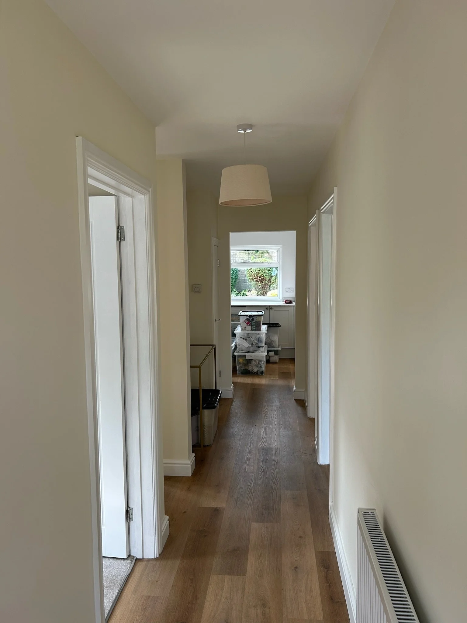 A hallway with light yellow walls, wooden flooring, and white door frames leading to a kitchen area with a window showing greenery outside. A hanging ceiling light fixture is present.