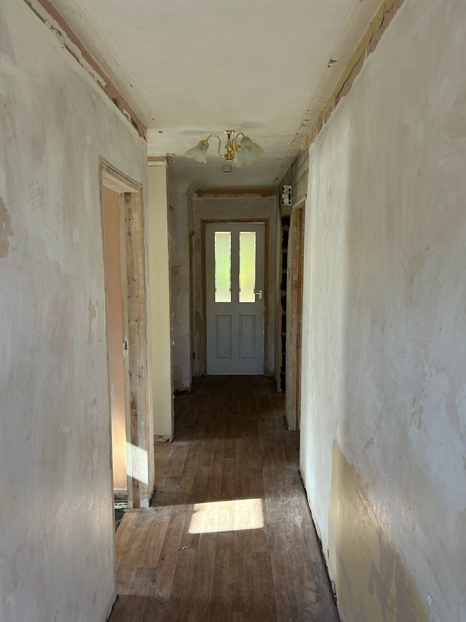 A hallway under renovation with unfinished walls and a wooden floor, leading to a door with a window at the end.