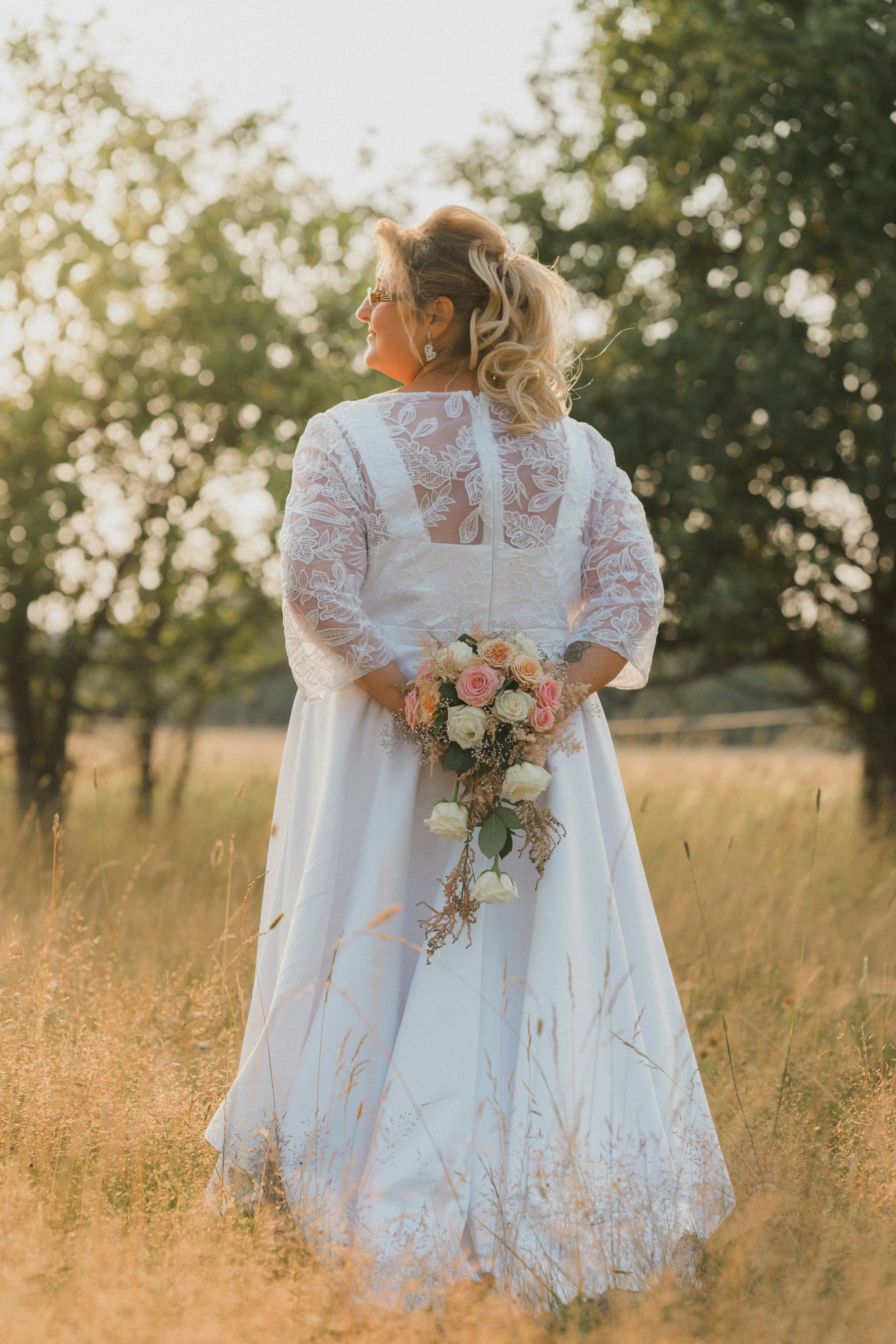 A bride in a white wedding dress holding a bouquet of pink and white roses, standing in a field with tall grass and trees in the background during sunset. Lincolnshire dress boutique Dress Me Up.