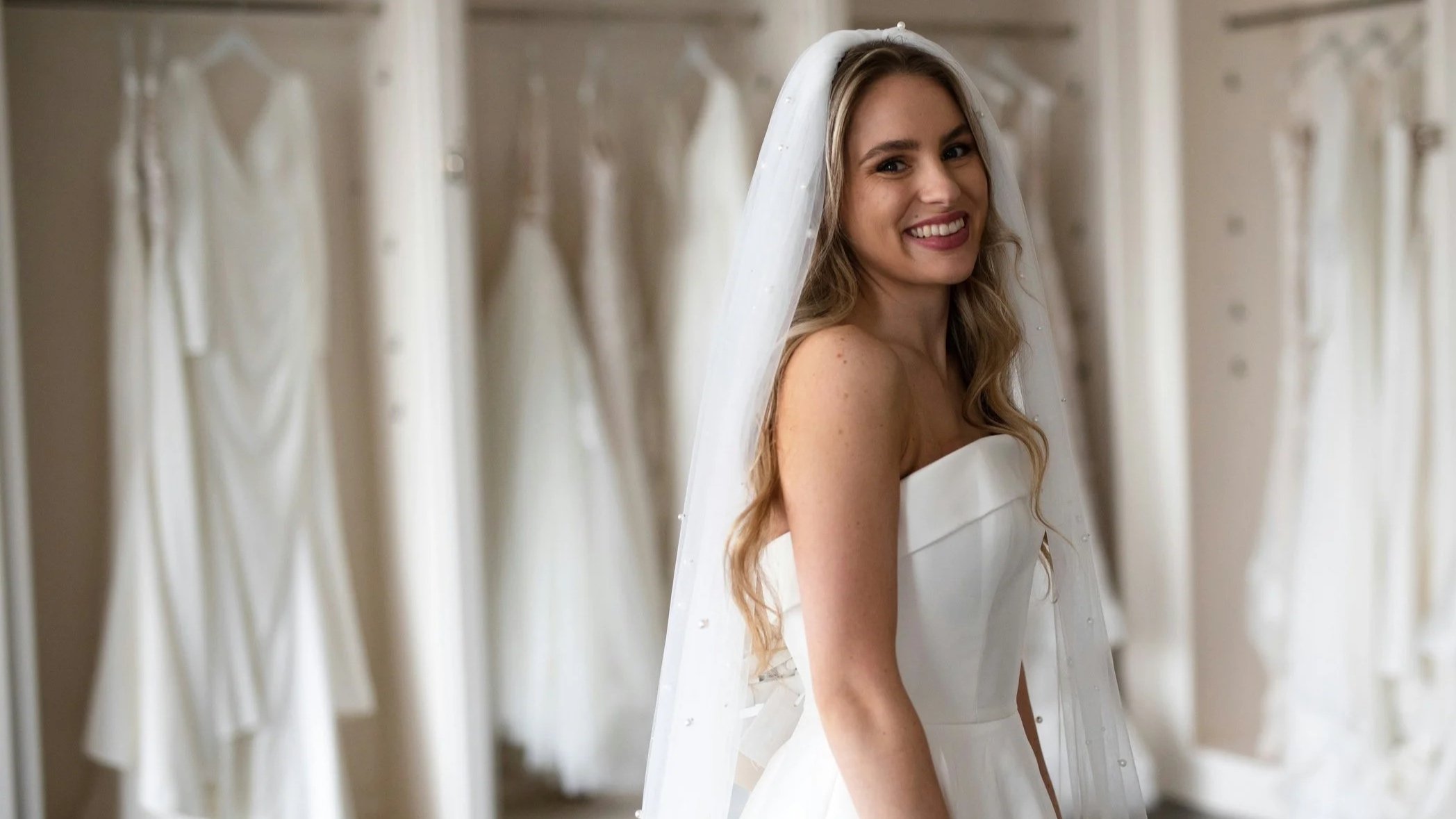 A bride in a white wedding dress smiling while trying on a wedding gown in a fitting room surrounded by wedding dresses hanging on racks.