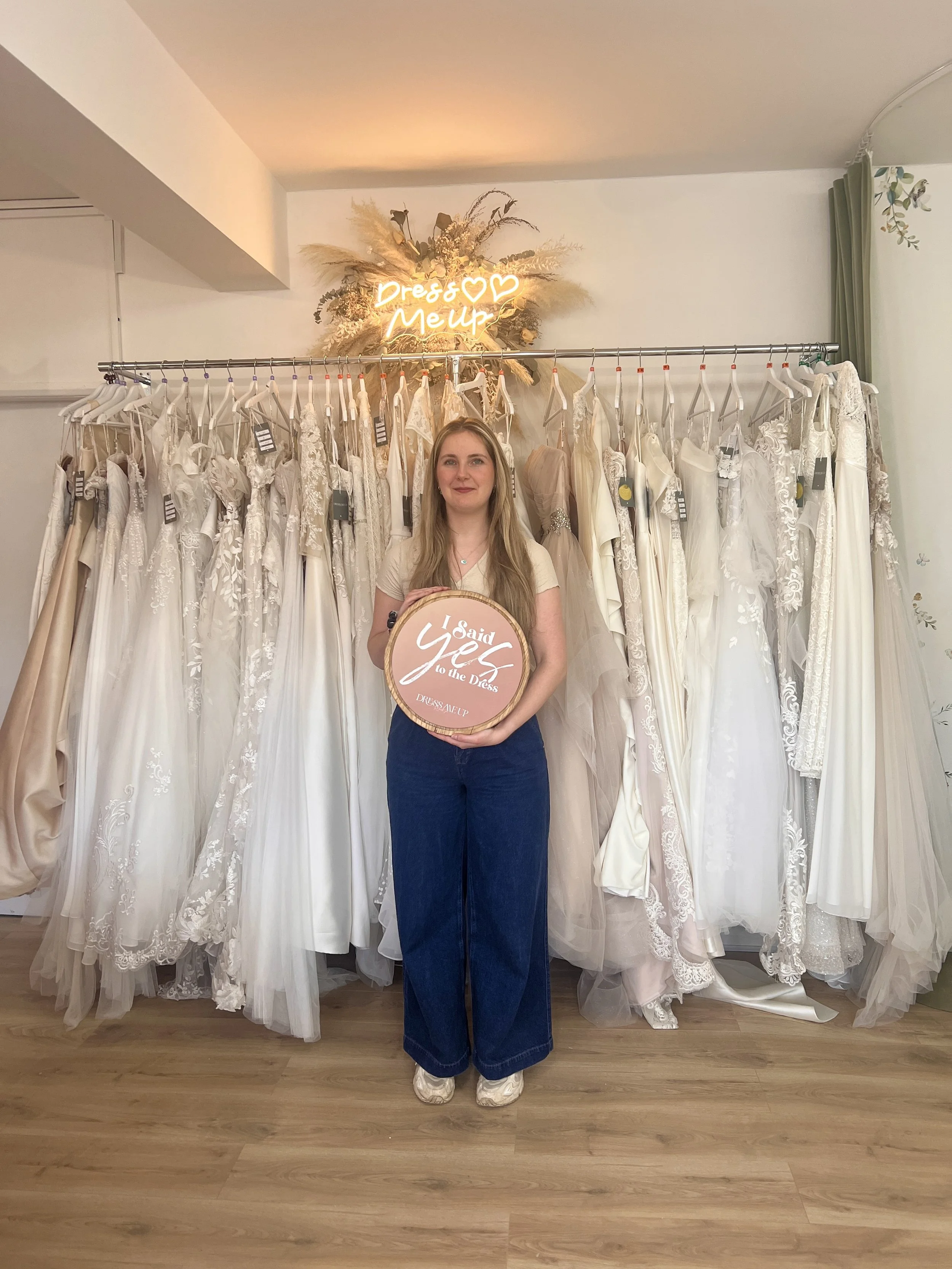 Woman standing in front of a rack of white wedding dresses, holding a circular sign that says 'I Said Yes to the Dress'. Behind her, there's a decorative sign with the words 'Dress & Make Up' illuminated in neon, surrounded by dried flowers and greenery.