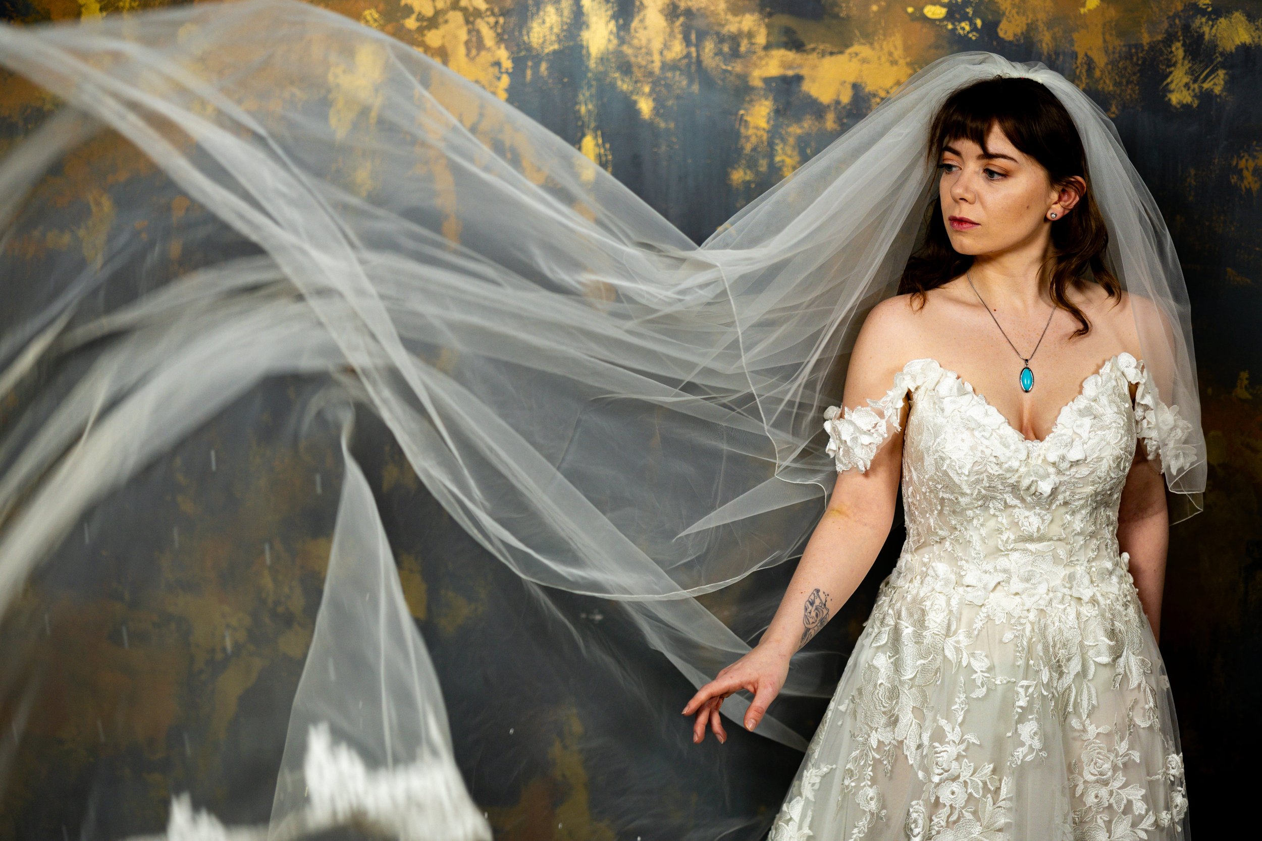 A woman in a wedding dress with lace details and off-the-shoulder sleeves, looking to her right. She is wearing a turquoise pendant necklace and has tattoos on her left arm. A long, flowing veil is draped over her head and flowing behind her, set against a dark, textured background.