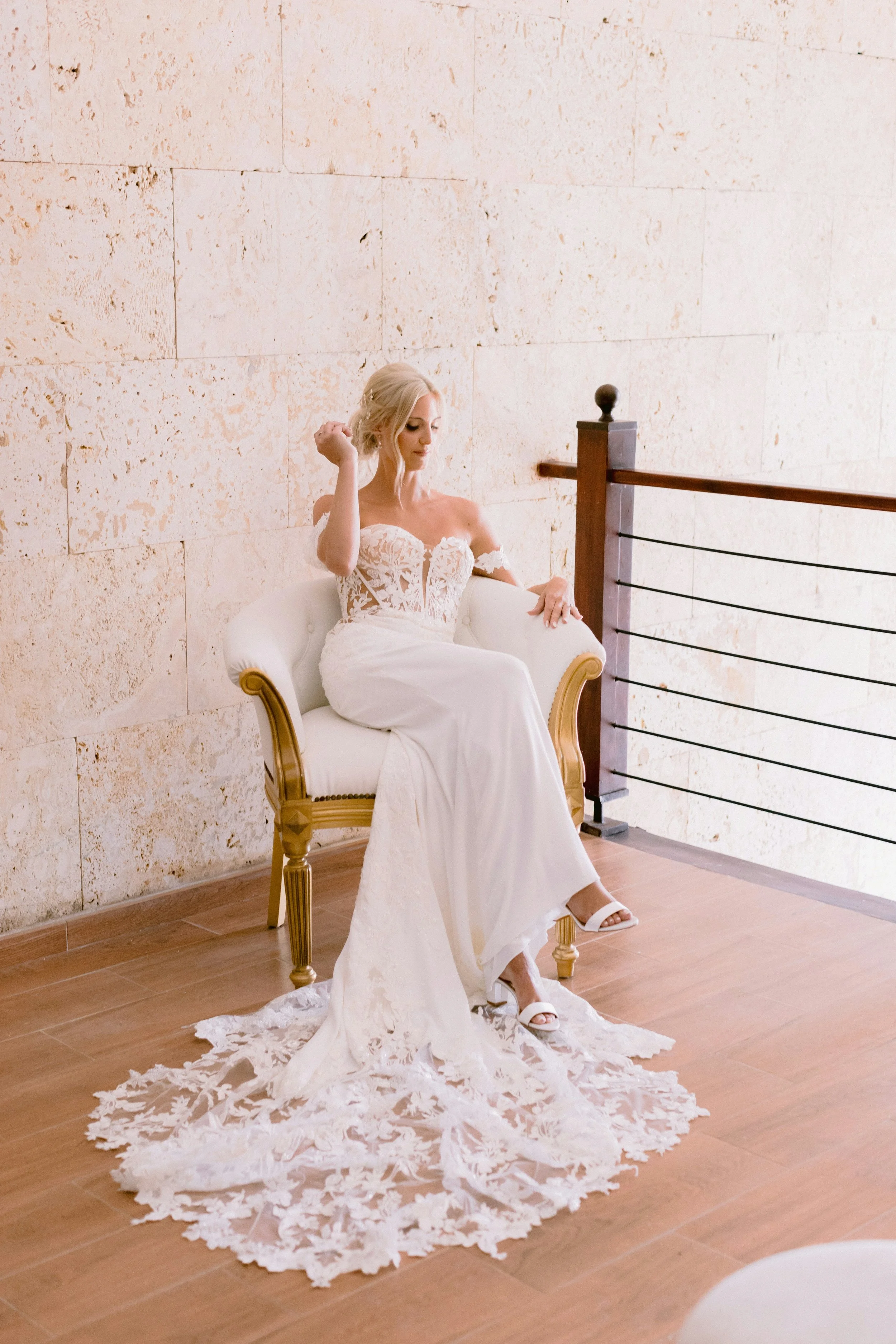 A woman with blonde hair in a wedding dress sitting on a white and gold cushioned bench, looking down, with her right arm raised near her head, on a wooden floor next to a stone wall and a wooden railing. Lincolnshire dress boutique Dress Me Up.