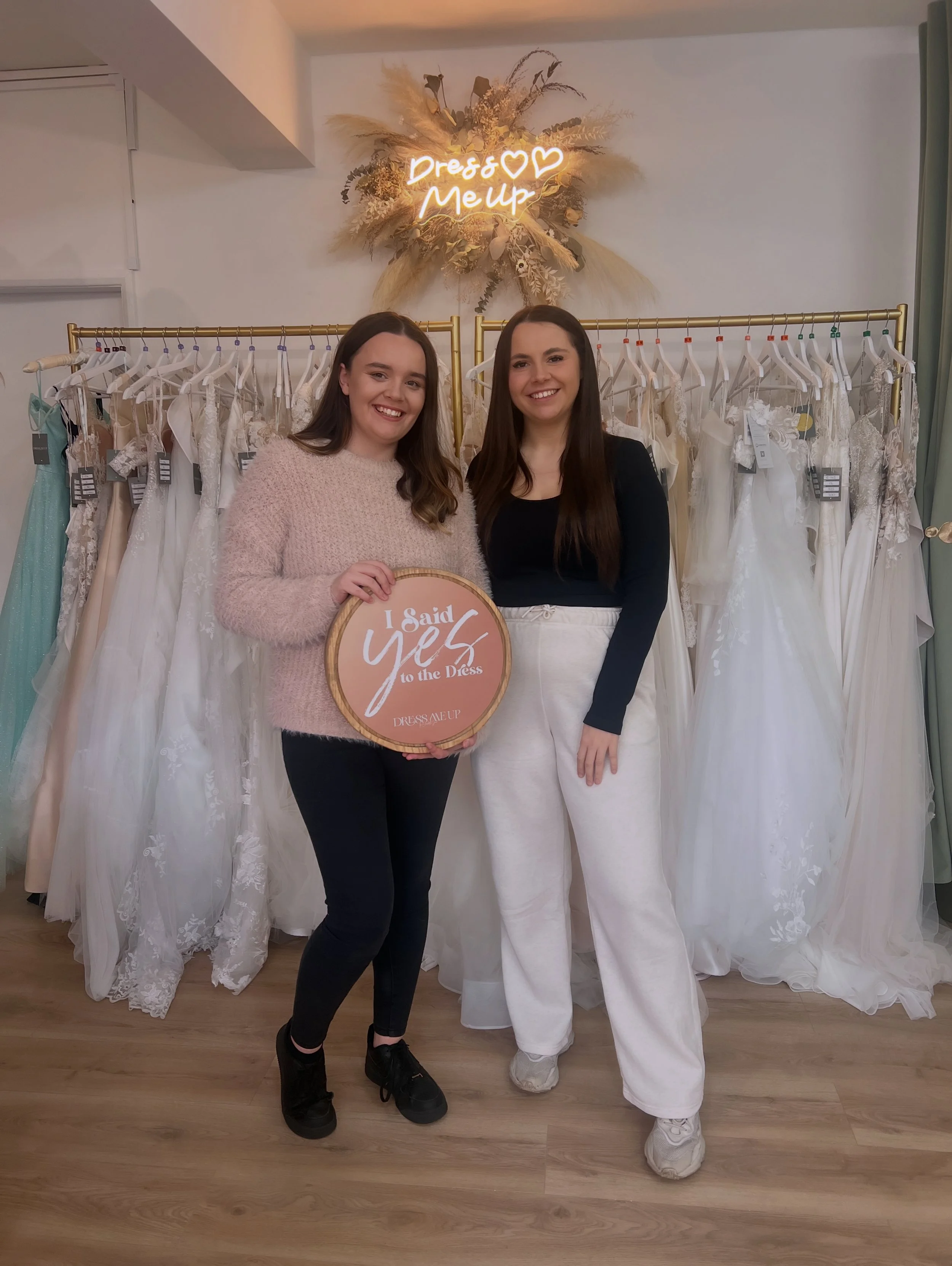 Two women standing in front of a clothing rack filled with wedding dresses, smiling for a photo. One woman holds a round sign that reads 'I Said Yes to the Dress.' Behind them, a decorative sign says 'Dress & Makeup' with a floral and pampas grass arrangement above.