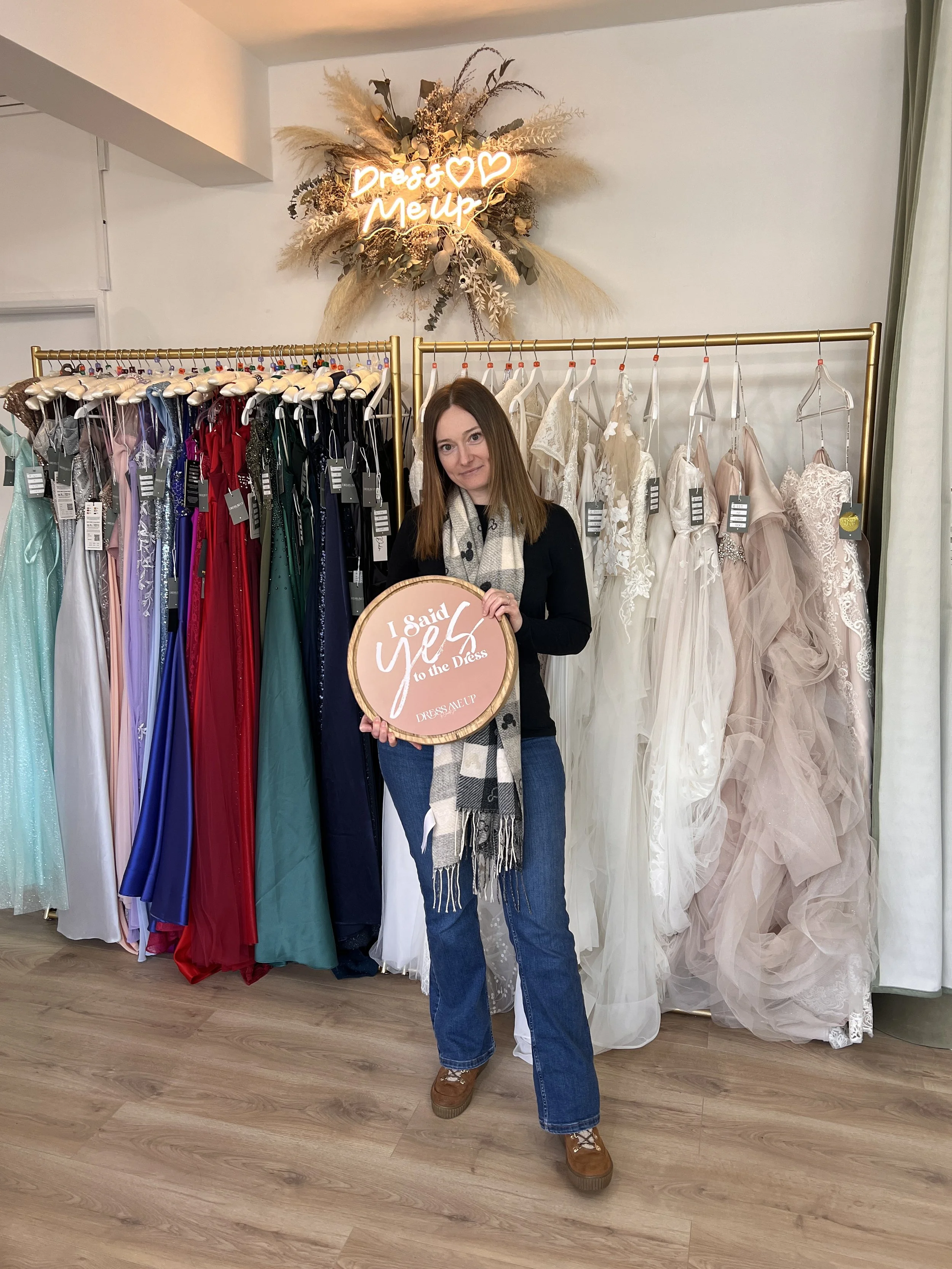 A woman with brown hair, wearing a black top, blue jeans, brown shoes, and a gray scarf with black hearts, stands in front of a rack of colorful and white dresses inside a boutique. She holds a round sign that says 'I Said Yes to the Dress.' On the wall behind her, there is a floral arrangement with a neon sign that says 'Dress & Me Up' with three hearts.