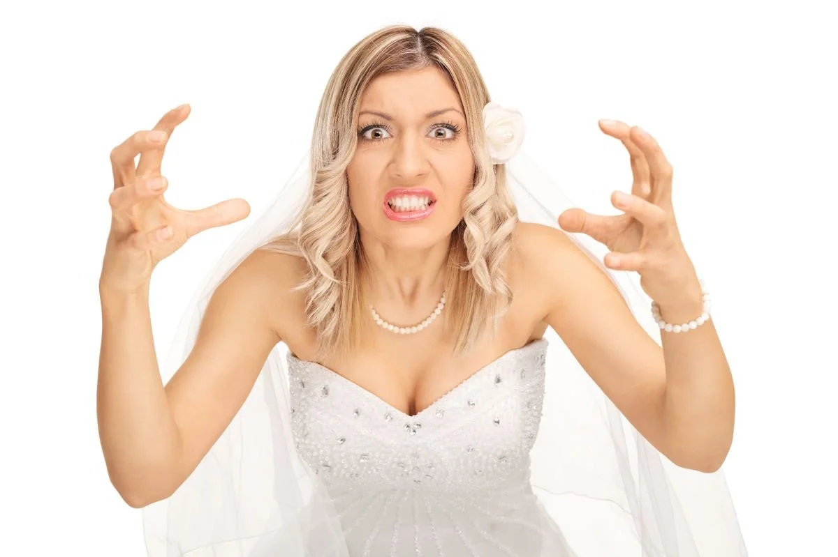 A woman dressed as a bride with an angry or frustrated expression, clenched hands, wearing a wedding dress with pearl jewelry, and a veil with a flower accessory in her blonde hair.