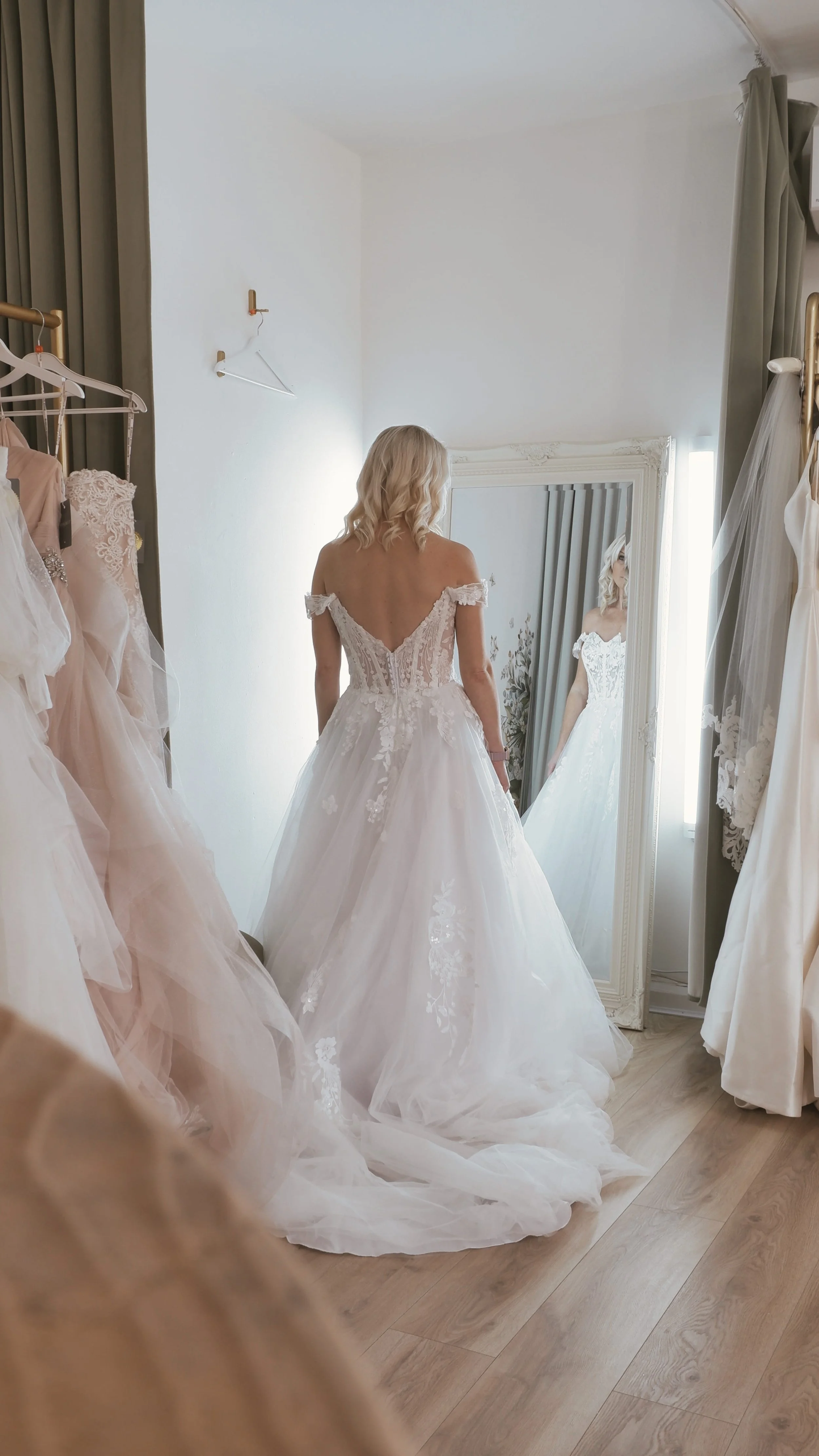 A woman trying on a wedding dress in a boutique, standing in front of a mirror. The dress is white with an off-the-shoulder design and lace details, and the boutique has several other wedding gowns hanging nearby.