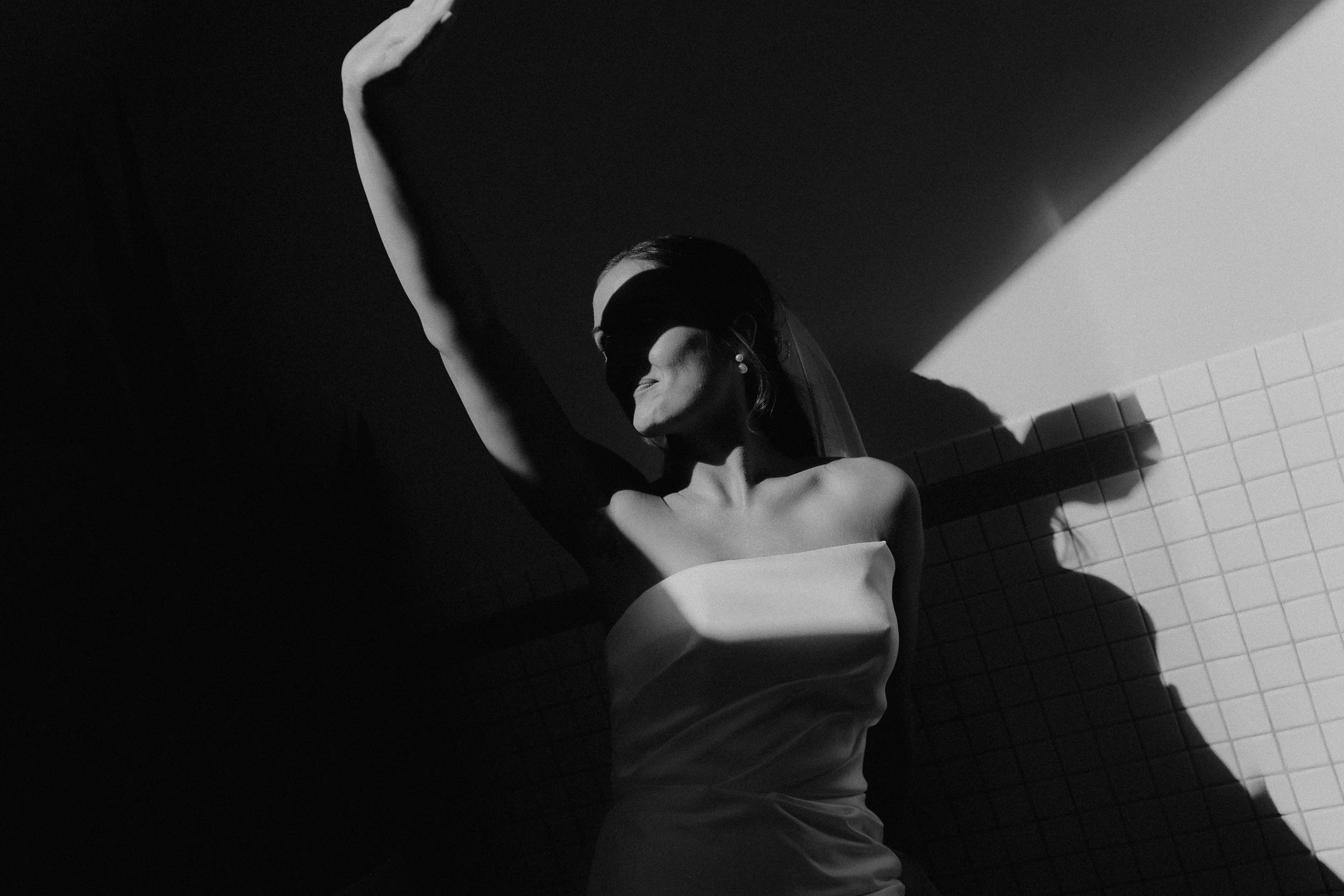 A woman in a strapless dress is standing against a tiled wall, with her arm raised. Shadows and light create a dramatic effect in this black and white photo.