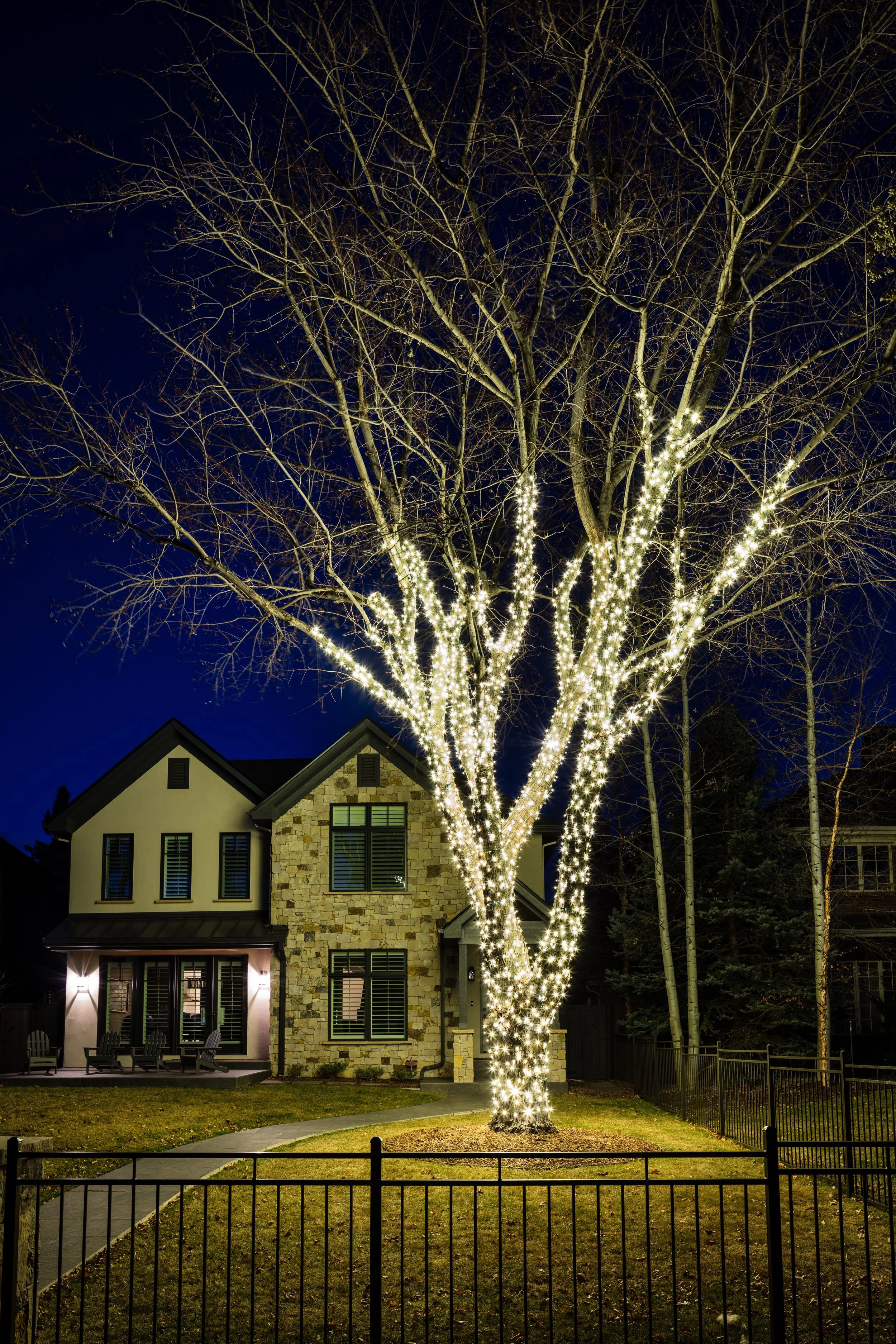 A large leafless tree decorated with white string lights stands in front of a two-story house at night. The house features stone and stucco exterior walls, several windows with shutters, and a small porch with Adirondack chairs. The yard is enclosed 