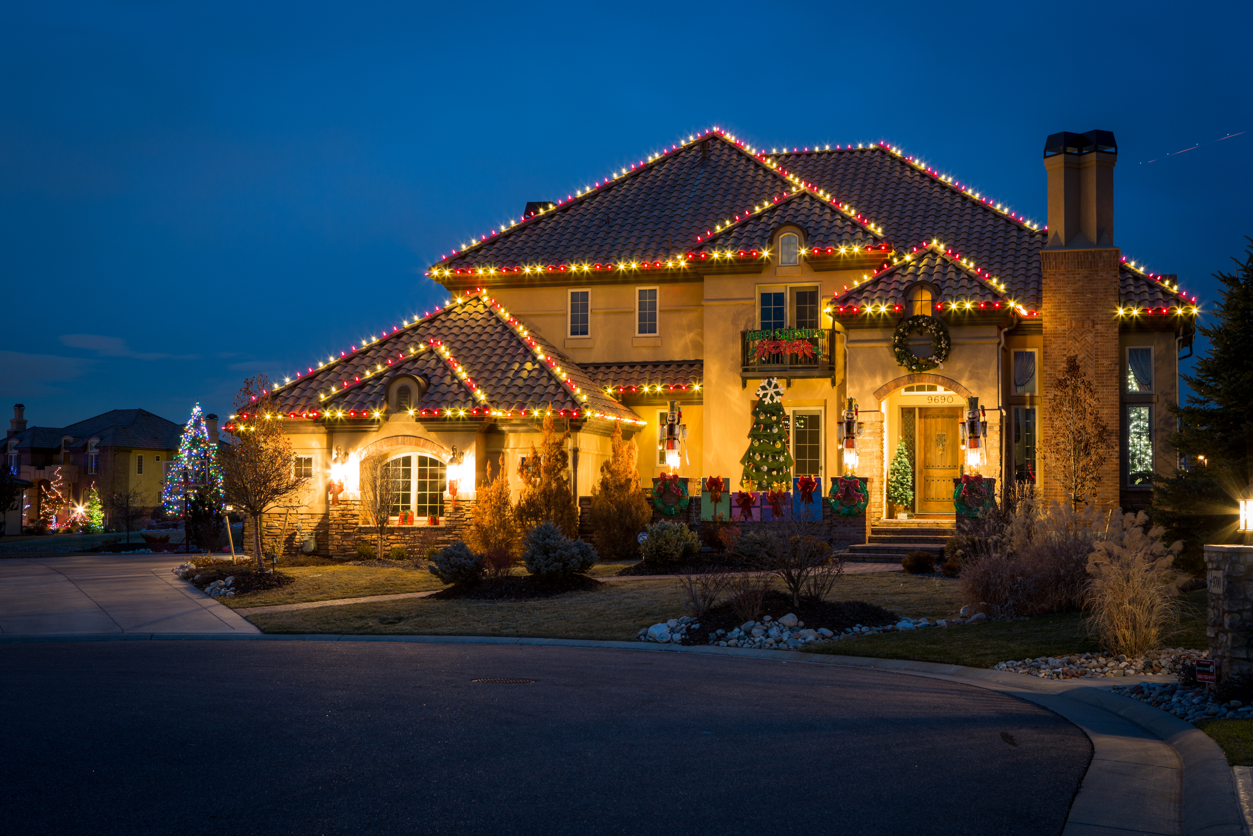 A large house decorated with Christmas lights, wreaths, and garlands during the evening. The house has a tiled roof, a chimney, and a front porch with wrapped gift boxes, a Christmas tree, and holiday decorations.
