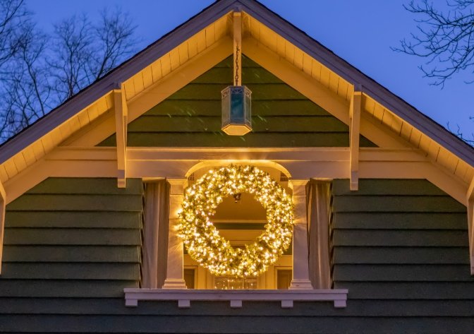 House with a gable roof decorated for Christmas, featuring a illuminated wreath in the window and hanging lantern outside.