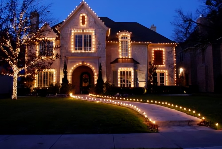 A large house decorated with Christmas lights at night. The house has a stone facade, multiple windows, and a curved pathway lined with small lights leading to the front door. There are additional lights outlining the roof and trees surrounding the h