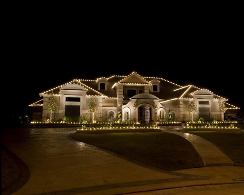 Large, elegant house illuminated with string lights at night, featuring a curved driveway and landscaped front yard.