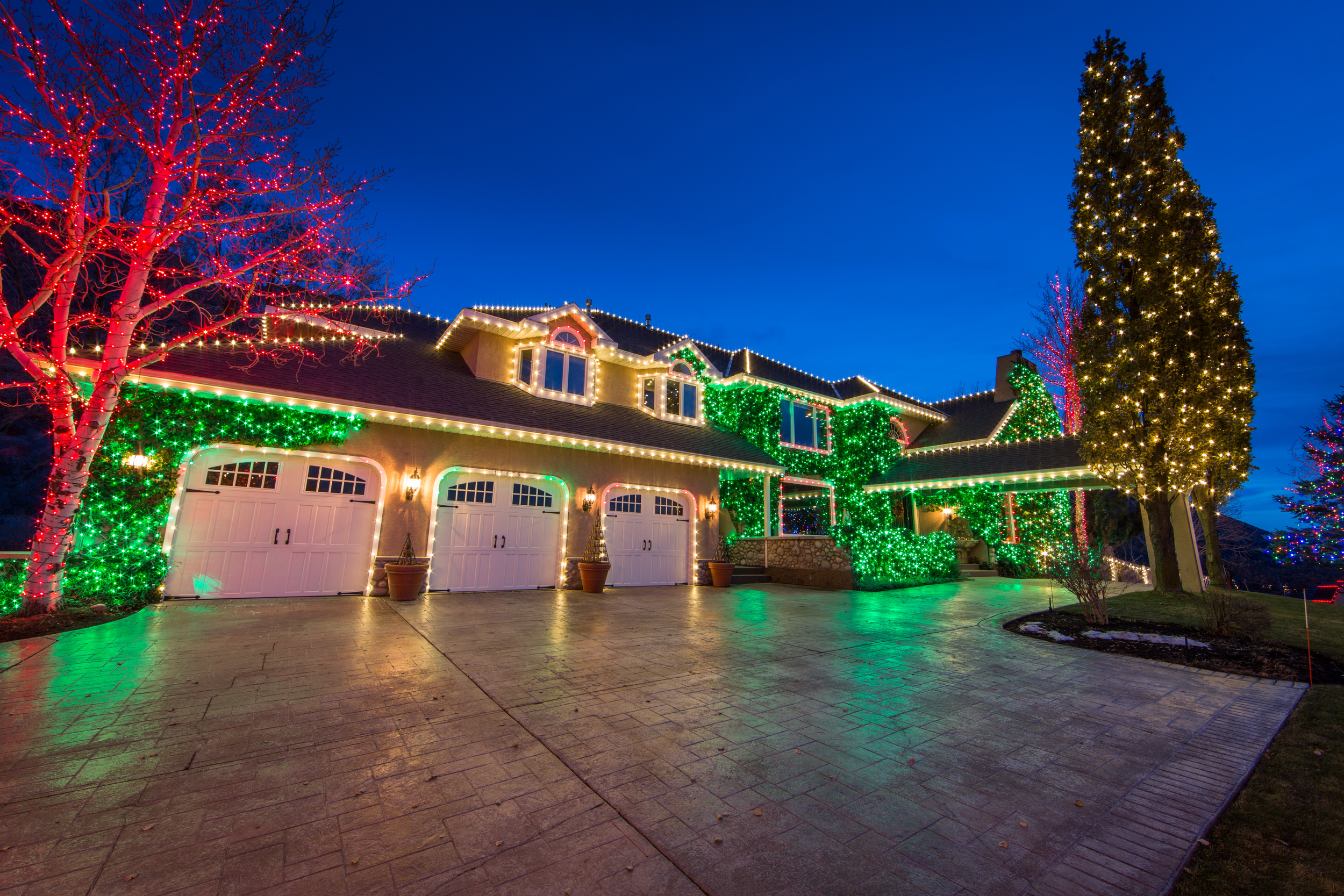 A house decorated with multicolored Christmas lights, with green, red, and white lights outlining the roof, windows, and trees. The driveway is illuminated with reflections of the lights, and the sky is dark blue.