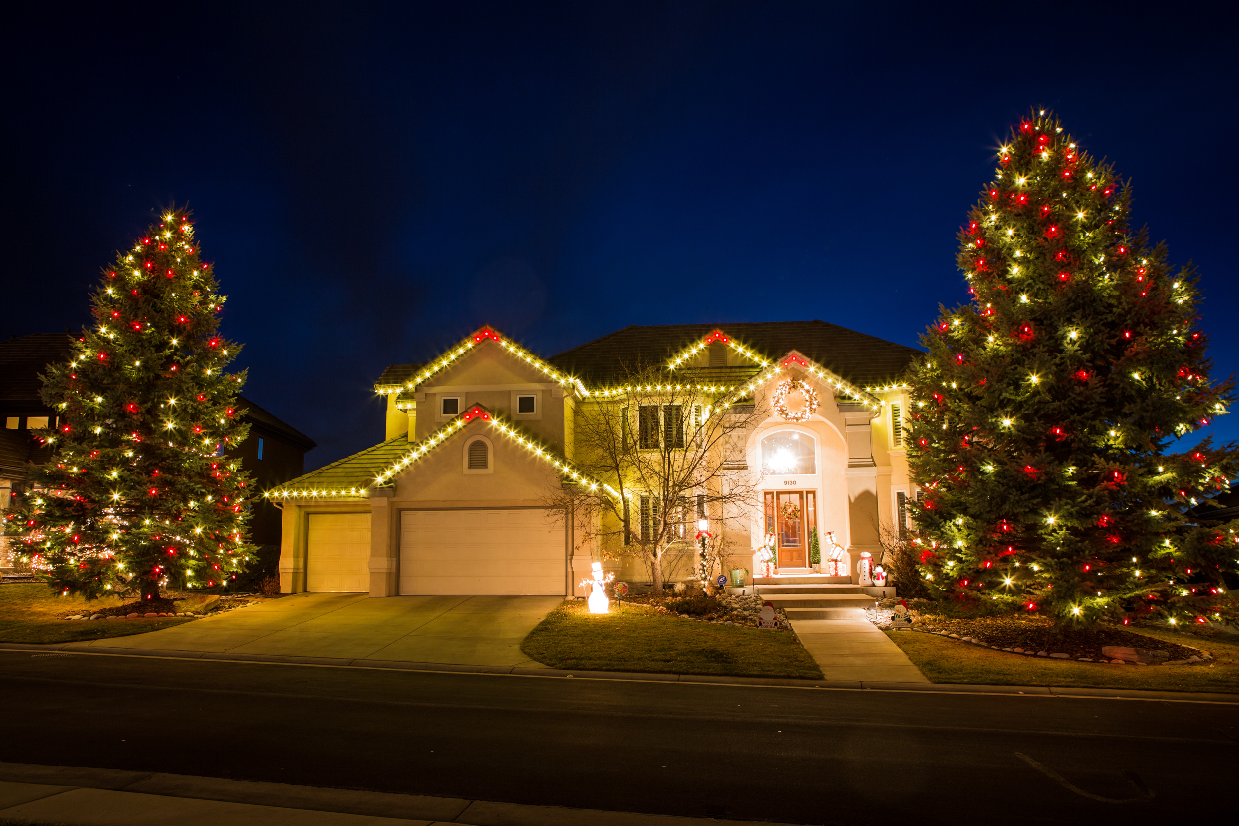 A house decorated with Christmas lights, including two large Christmas trees with red and yellow lights, and holiday-themed decorations on the front porch. It's nighttime with a dark blue sky.