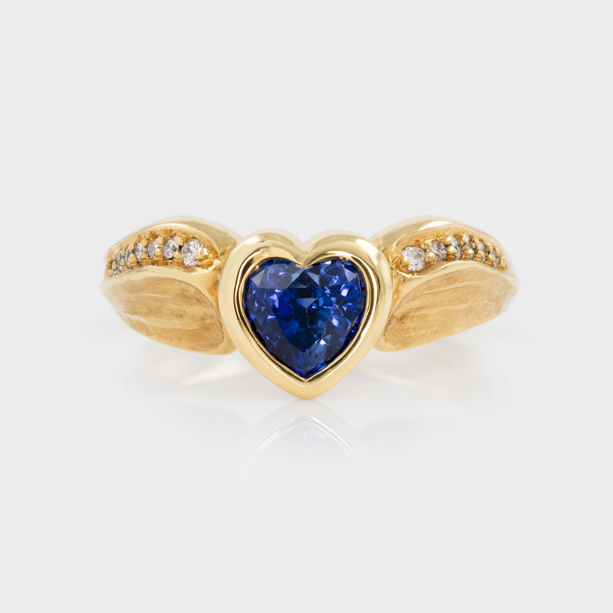 Make a SUPER gesture for Valetine's Day and surprise your loved one with this beautiful 18 karat yellow royal gold ring designed by Michael Endlich and featuring his signature redwood bark texture. The ring centers a heart shaped blue Sapphire, flank