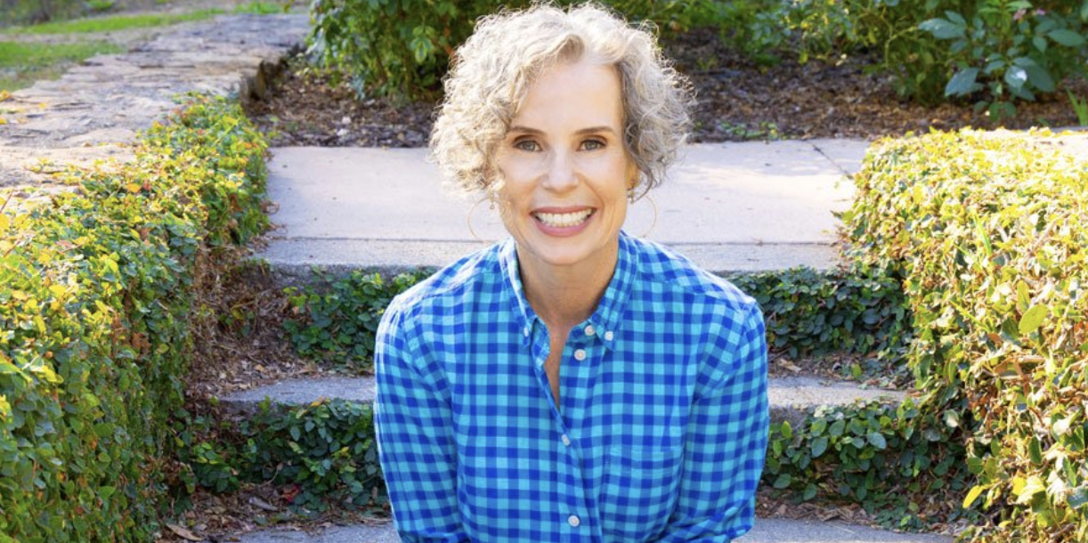 A smiling woman with short curly gray hair, wearing a blue checkered shirt, sitting outdoors on steps with green bushes and plants in the background.