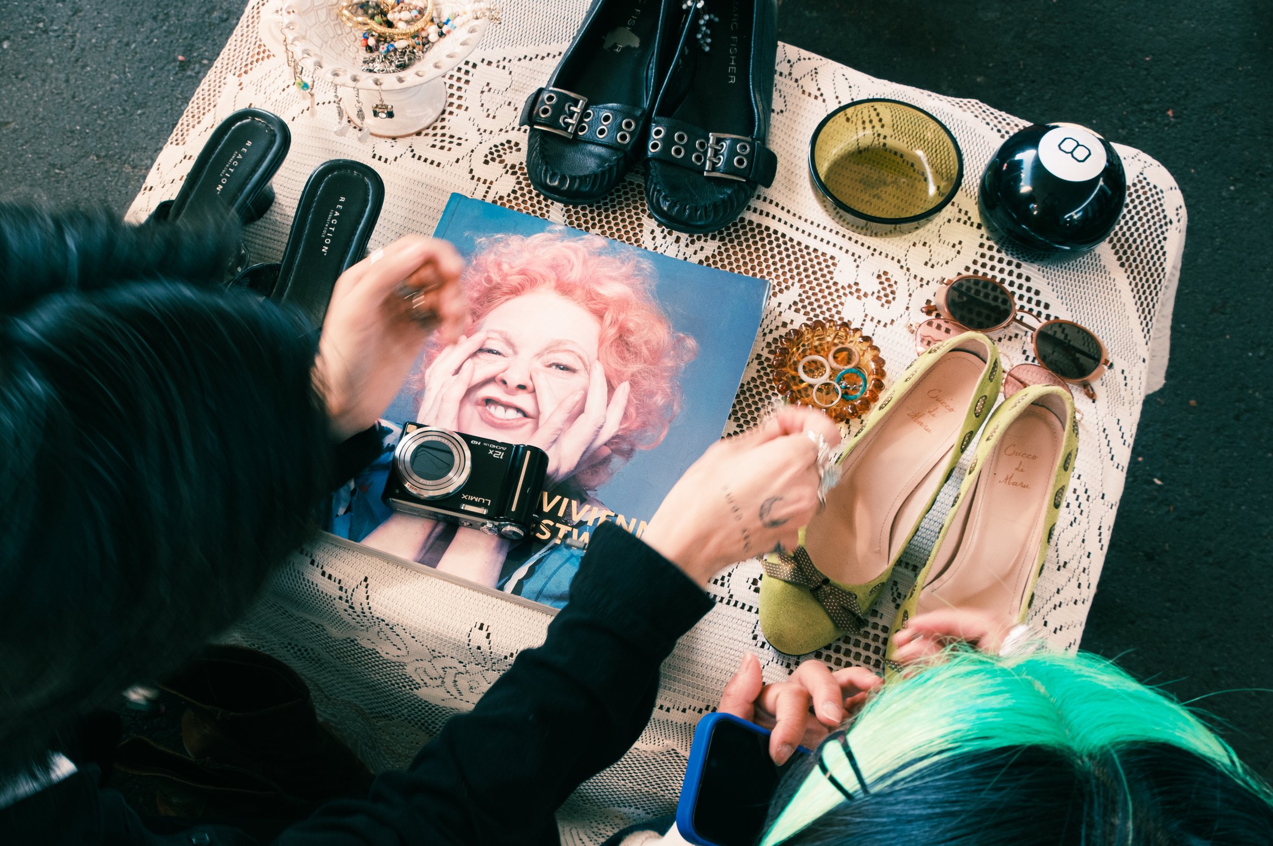 A person with long black hair and tattoos on their hands is browsing jewelry and accessories on a table at an indoor market. Items include sandals, sunglasses, bracelets, a camera, and a magazine featuring a woman with pink curly hair.