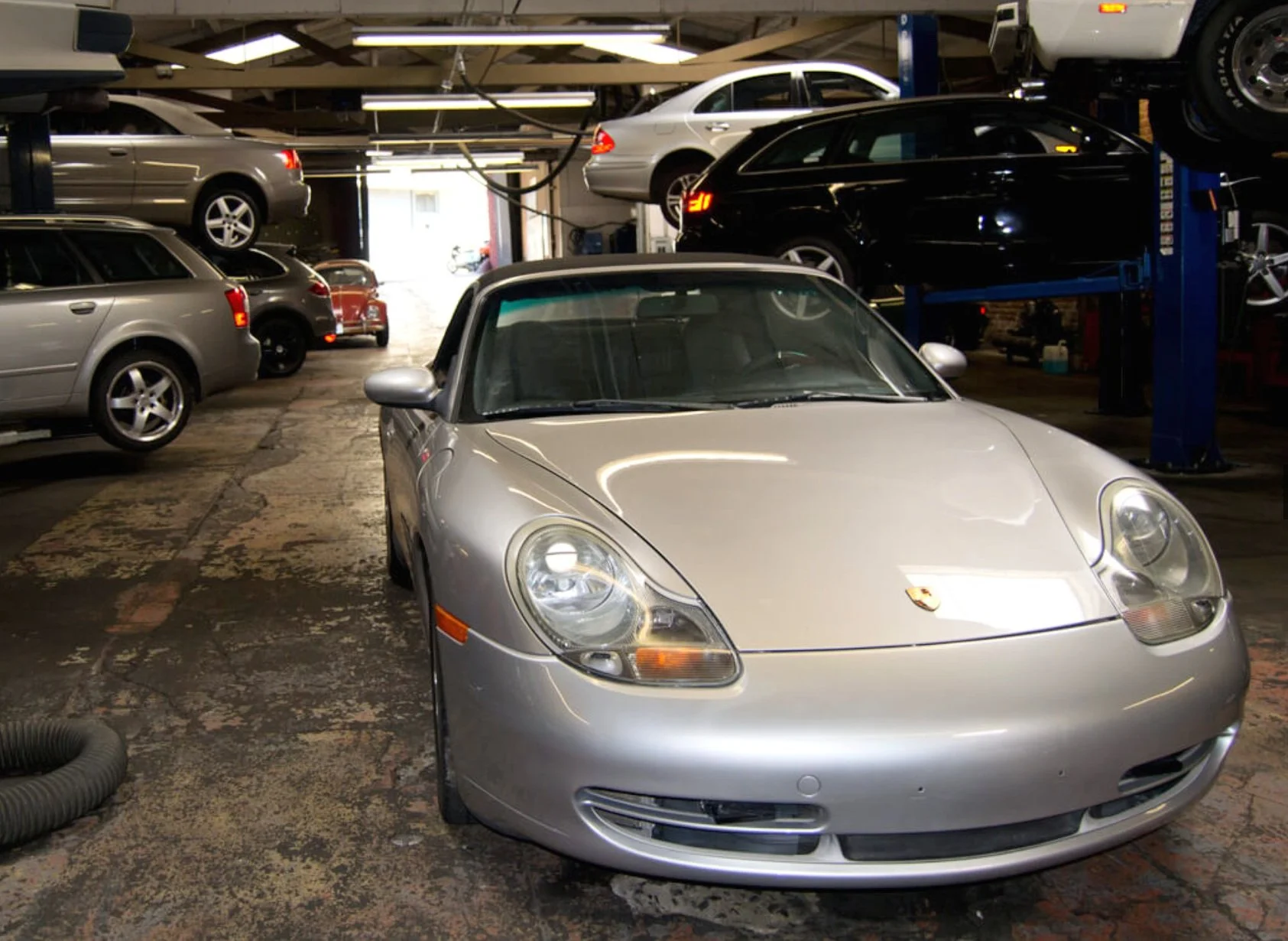 Silver Porsche sports car in an automotive repair shop with multiple cars and a car lift in the background.