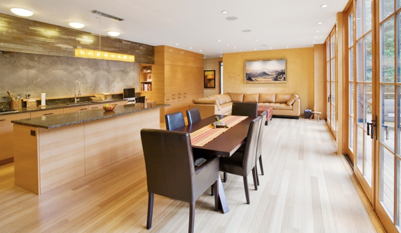 Open-concept living and dining area with a kitchen island, a dining table with six chairs, and a beige sectional sofa, featuring large windows with wooden frames allowing natural light.