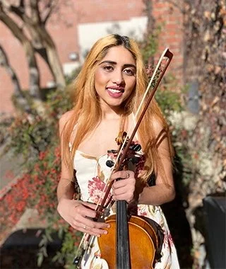Young woman with blonde hair holding a violin and bow outdoors in a park.