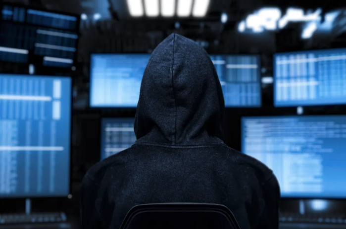 Person with a hoodie sitting at a computer station with multiple monitors displaying code or data.