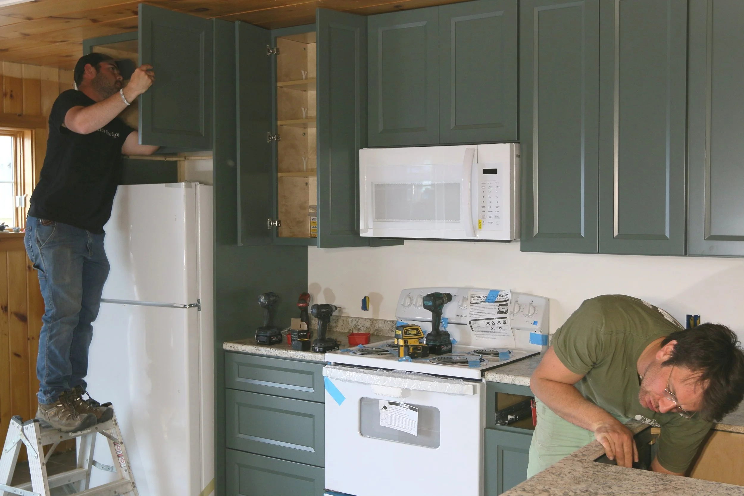 Two men working in a kitchen, installing cabinets and appliances. One man is on a ladder reaching into a cabinet above a refrigerator, and the other is crouched at the counter working on a cabinet.