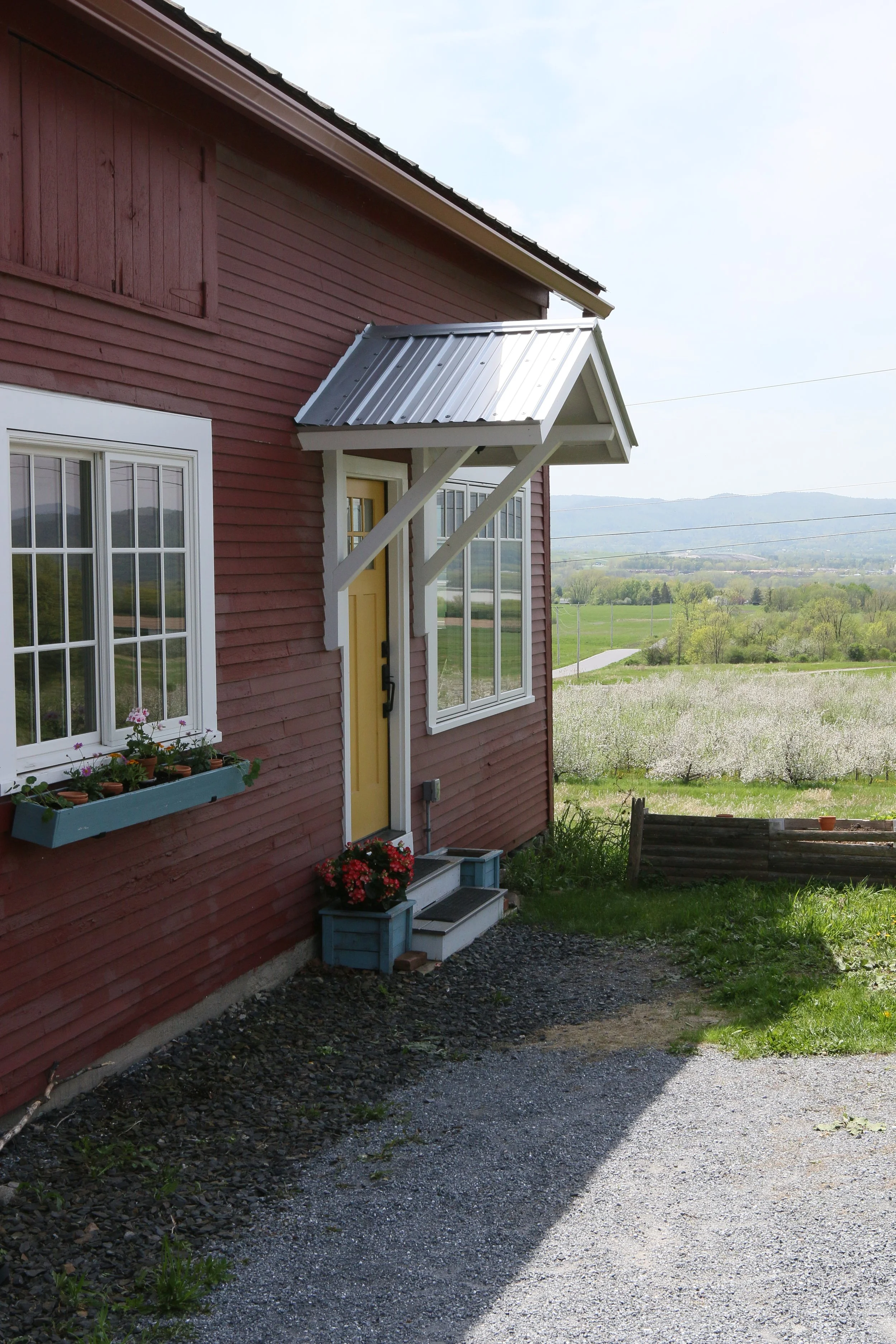 Red house with a yellow door, white-framed windows with flower boxes, a small set of stairs with potted flowers, and a metal awning over the door, overlooking a farm landscape with blooming trees and hills in the distance.