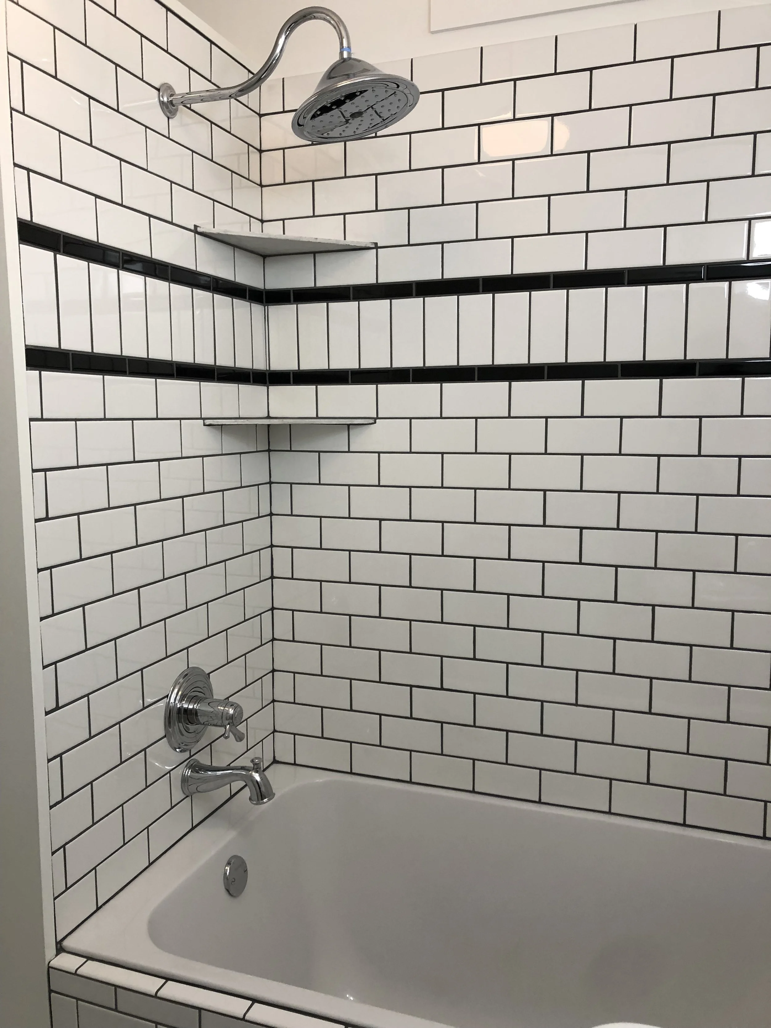 White tiled bathroom shower with black horizontal stripe tiles, a rain shower head, a single handle faucet, a soap dish, and corner shelves.