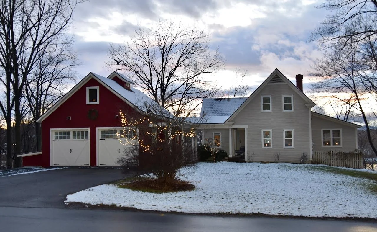A house with beige siding and a separate red garage with white doors, both with snow on their roofs, in a winter setting with leafless trees and a cloudy sky at dusk.