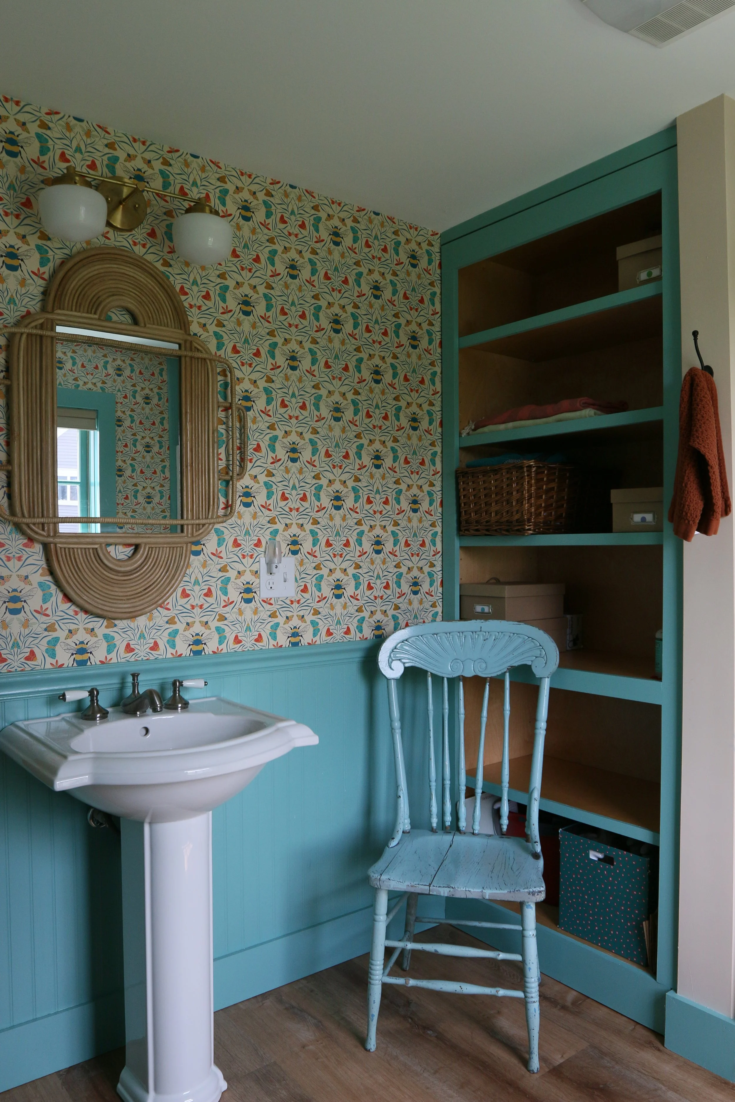 A vintage-themed bathroom with a small pedestal sink, a wooden chair painted light blue, a patterned wallpaper with bees and flowers, a square wall mirror with a rattan frame, a teal built-in shelf organizer, and a brass wall sconce with two white gl