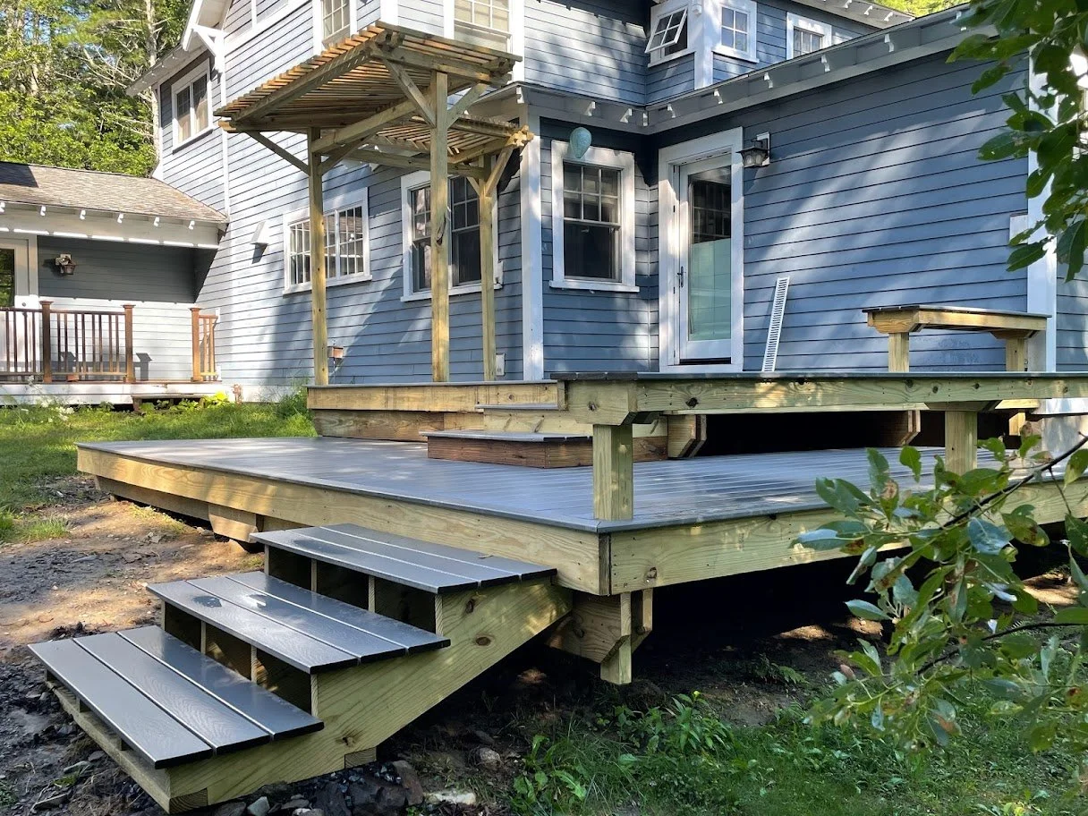 Newly built wooden deck attached to a blue house with stairs leading down to ground level, surrounded by grass and trees.